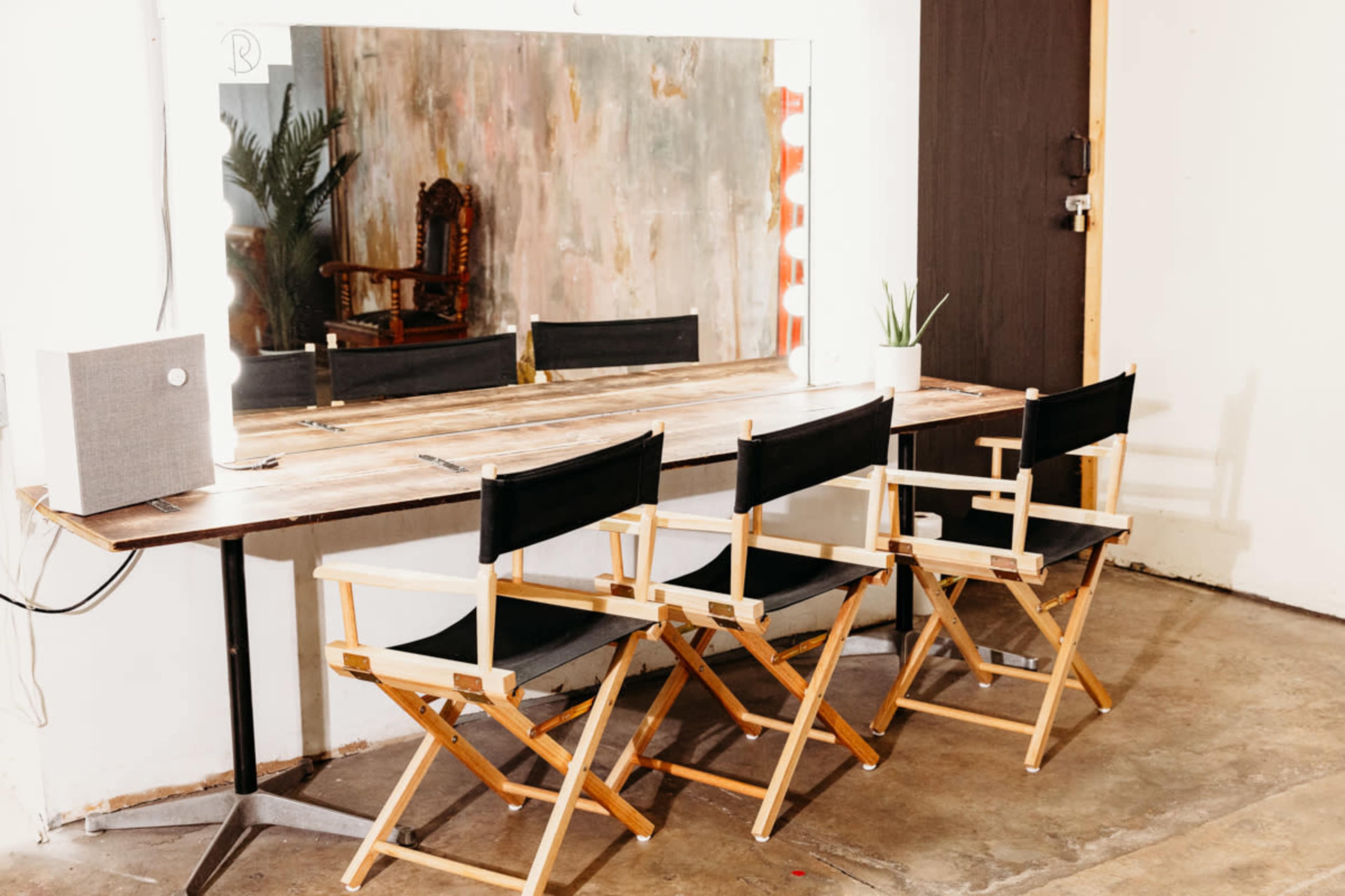 A wooden table with three director's chairs is positioned in front of a large mirror in a minimalist interior space.