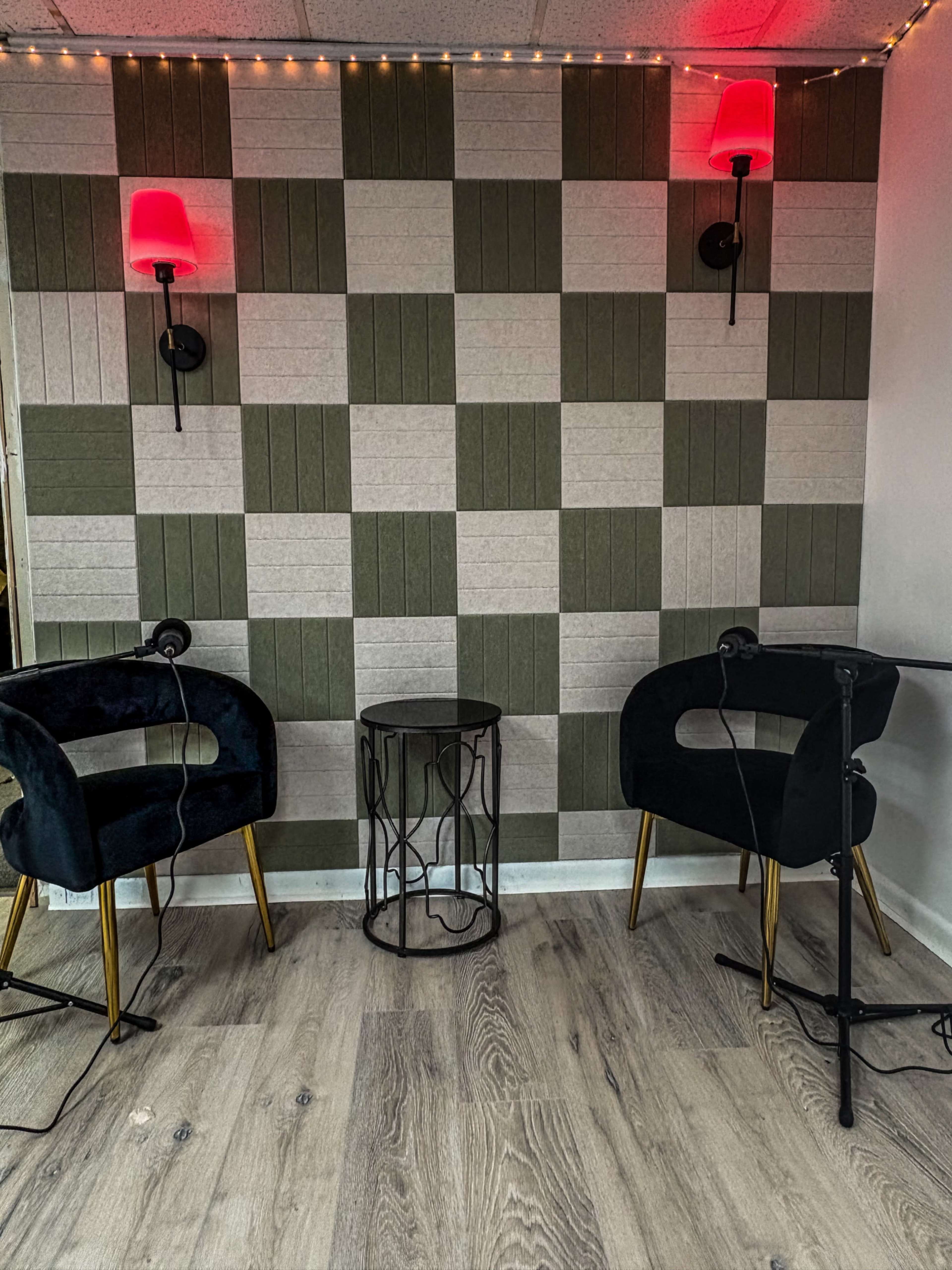 The image shows a cozy corner with two black velvet chairs, a small metallic side table, and a checkered wall design featuring alternating light and dark panels, illuminated by red wall sconces.
