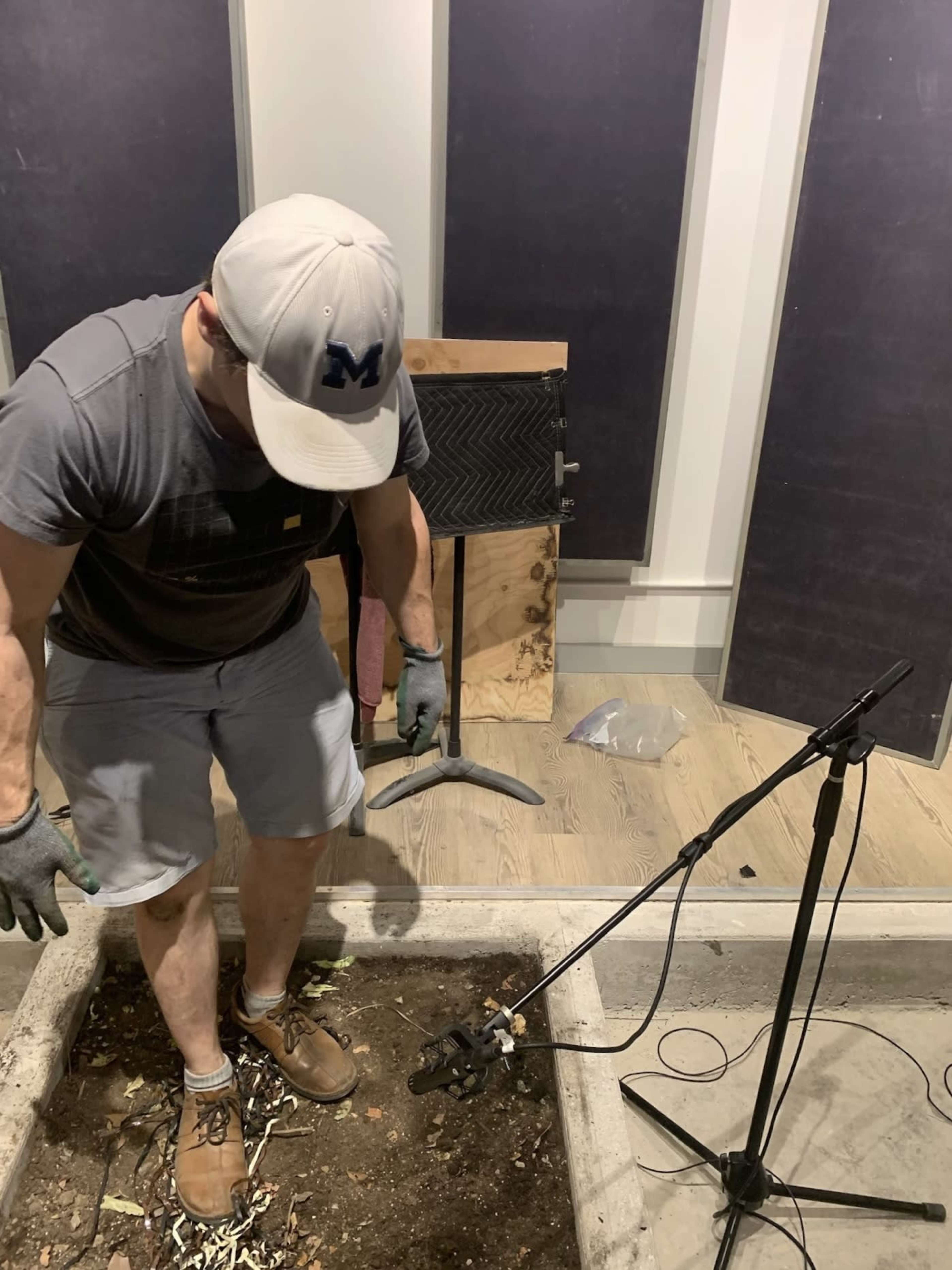 A person in a gray t-shirt and baseball cap stands in a planting bed, using a microphone stand while digging in the dirt.