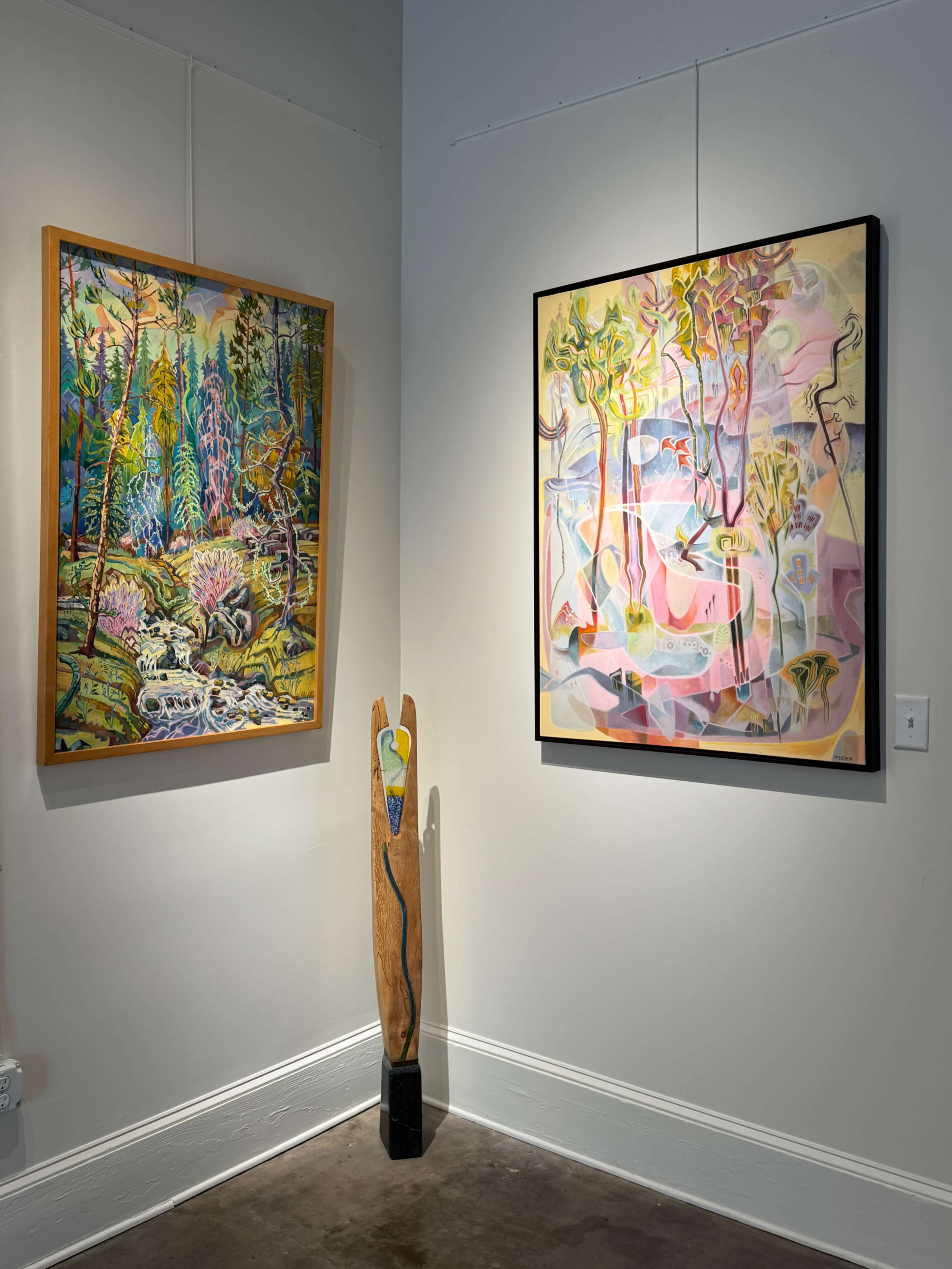 The image shows an art gallery interior with two colorful paintings on the wall and a wooden sculpture positioned in the corner.