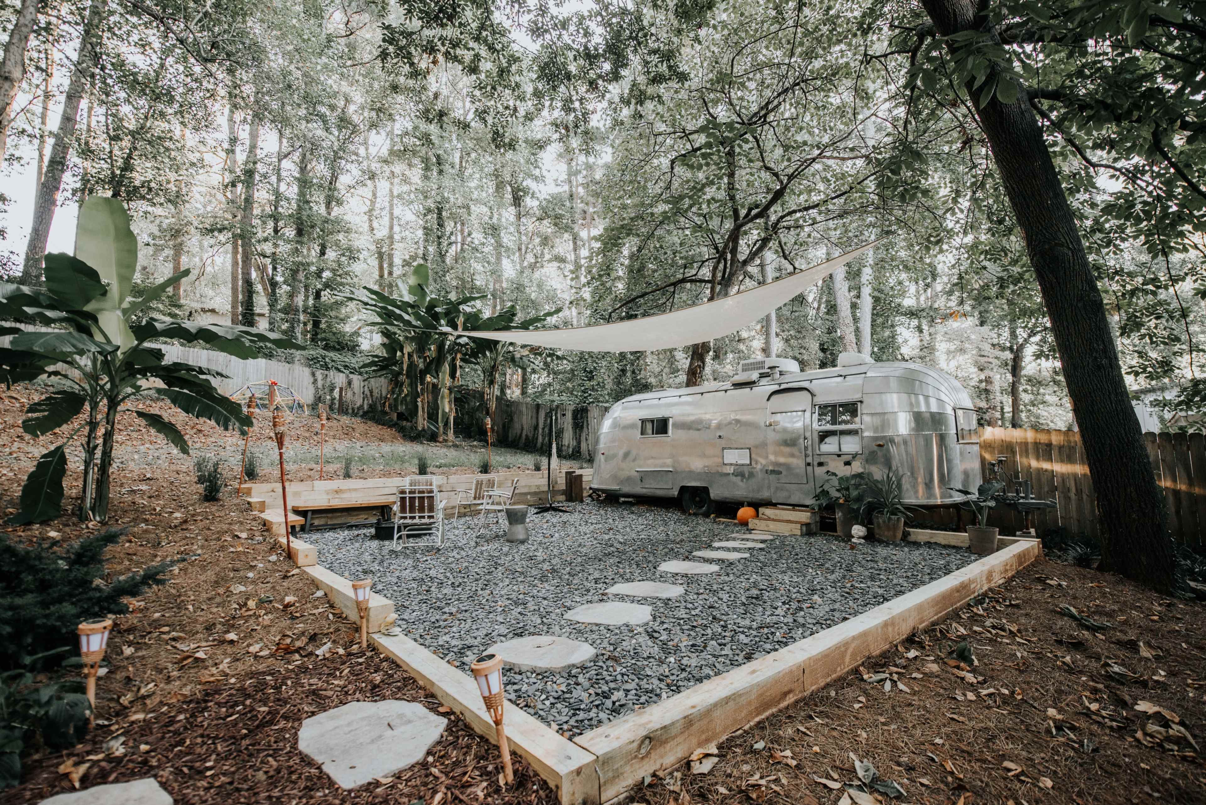 A vintage Airstream trailer is parked in a landscaped area with gravel, stone steps, wooden seating, and shade provided by a canopy in a forested setting.