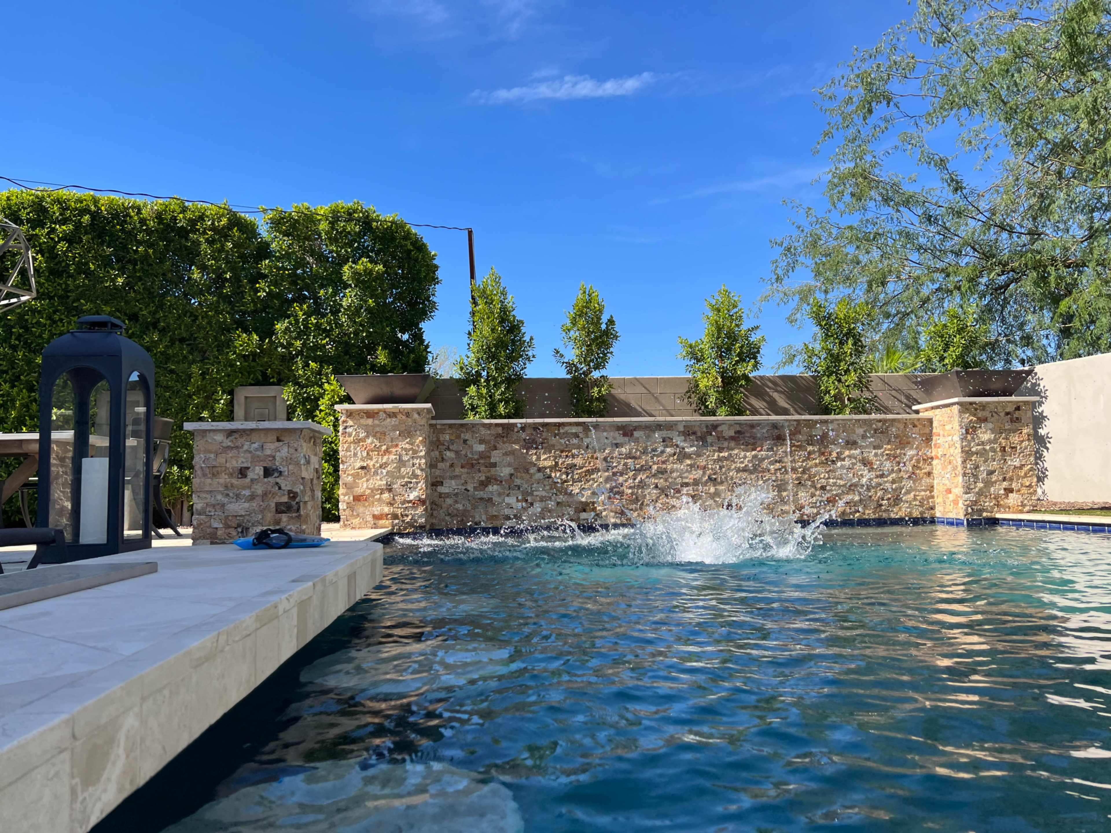 The image shows a clear swimming pool with a stone wall and greenery in the background under a blue sky.