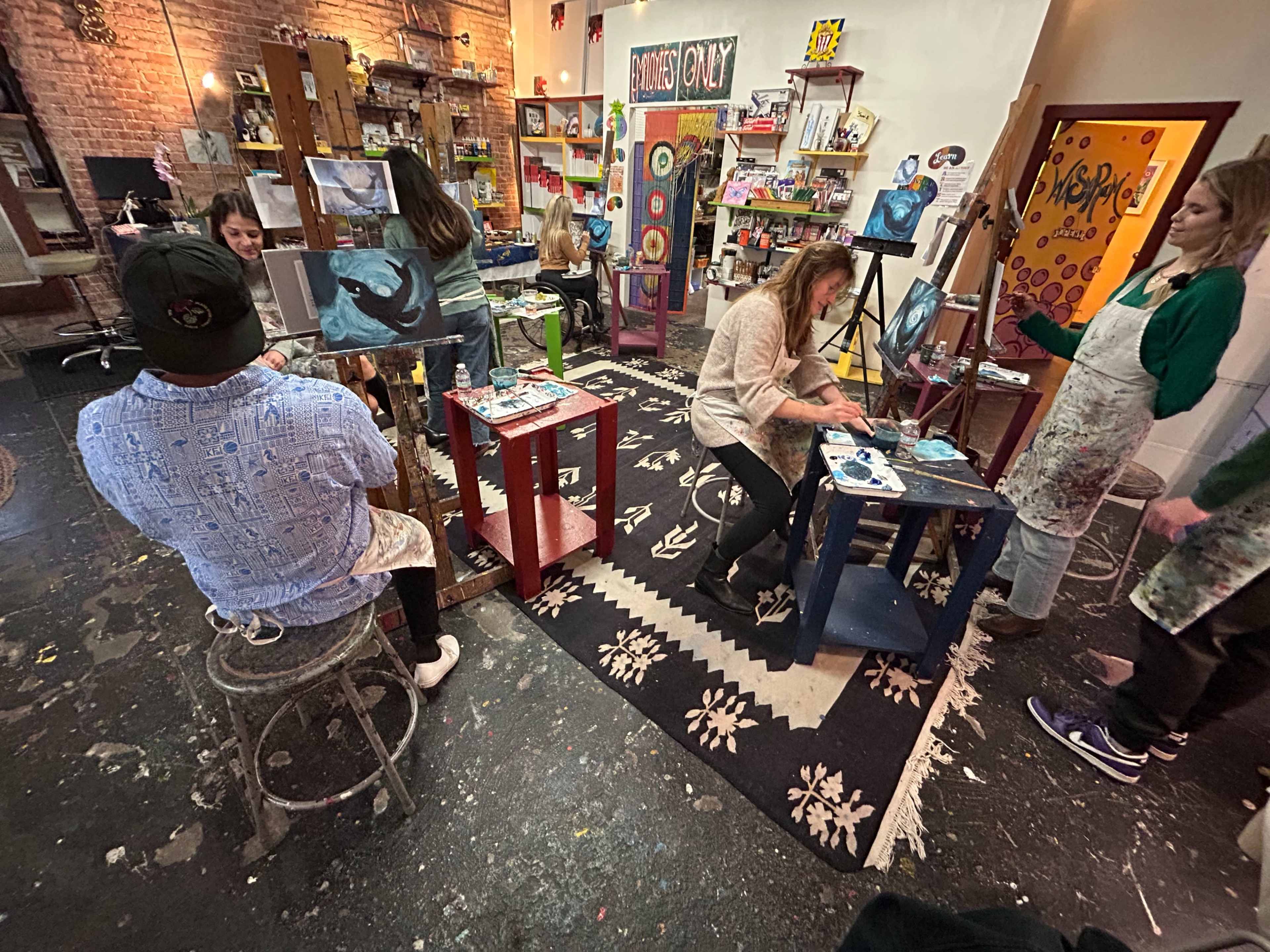 A group of people is painting on easels in a colorful art studio with various artworks displayed on the walls.