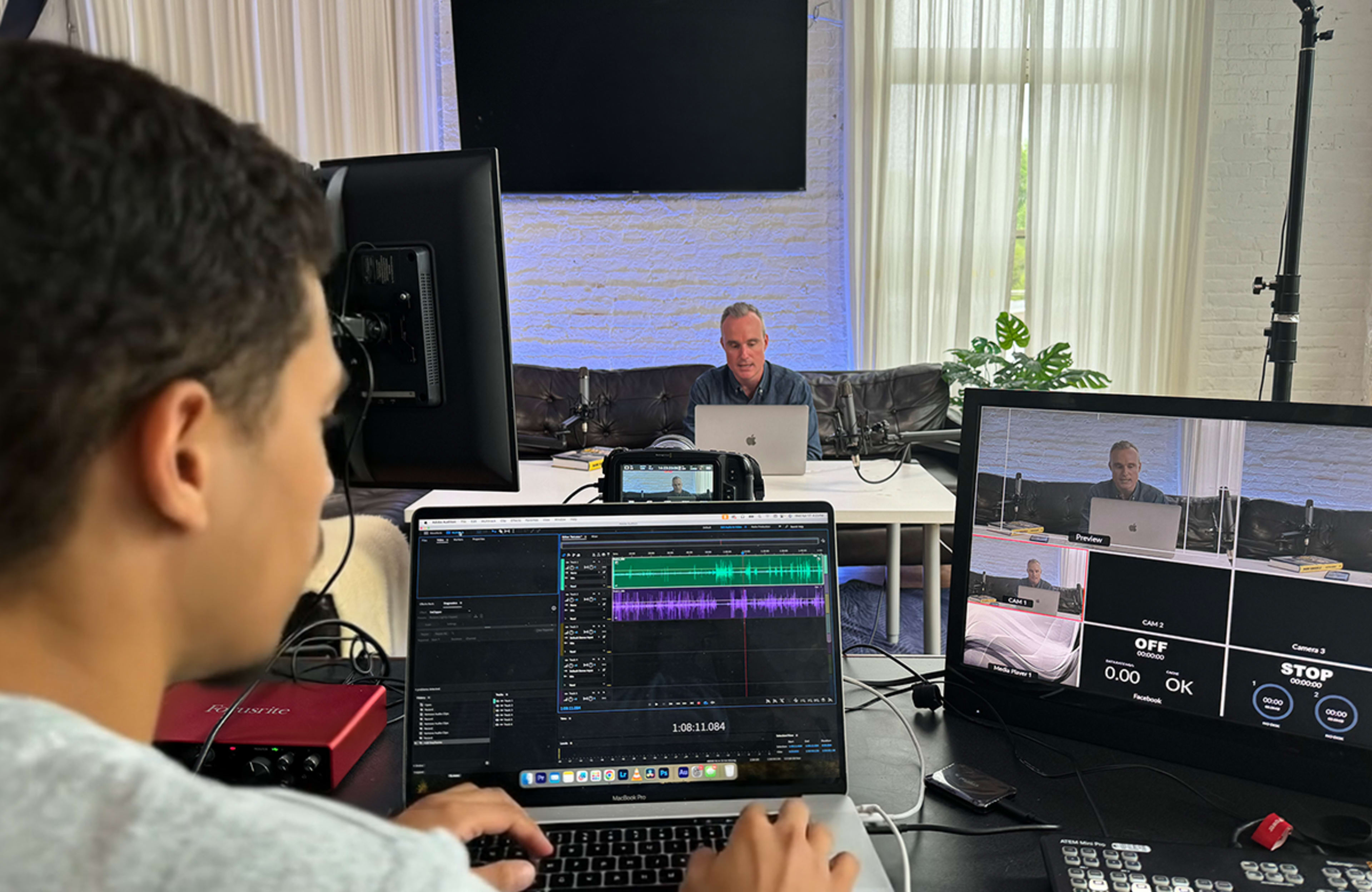 A person is editing audio on a laptop while another individual is seated at a table, visible on a monitor during a video recording session.