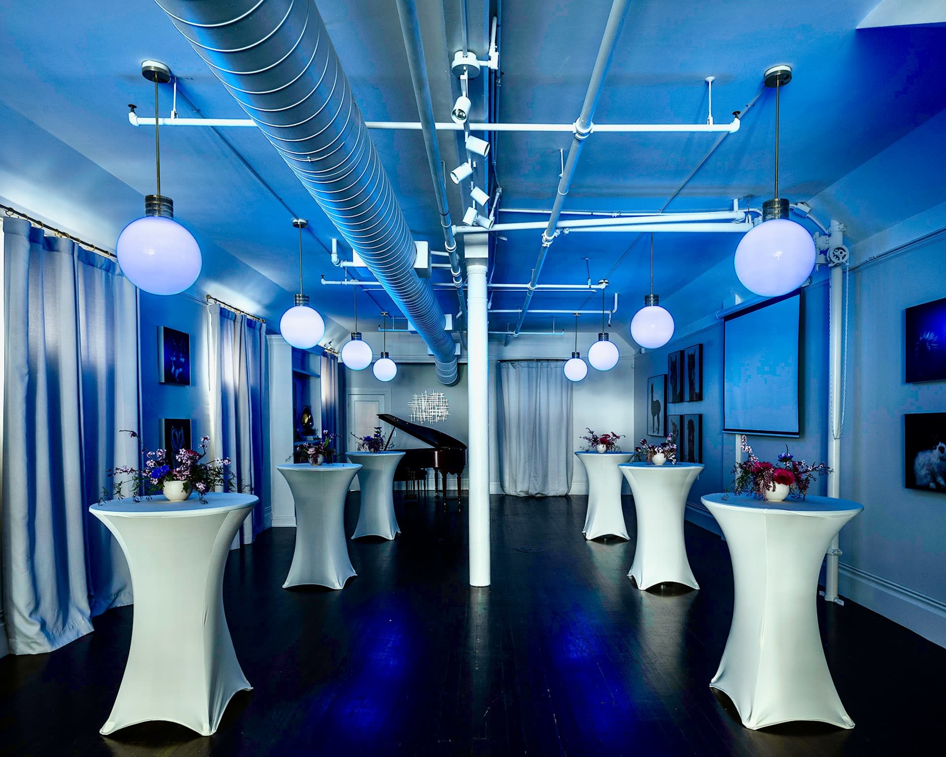 A modern event space features six white cocktail tables surrounded by blue lighting and decorative floral arrangements.