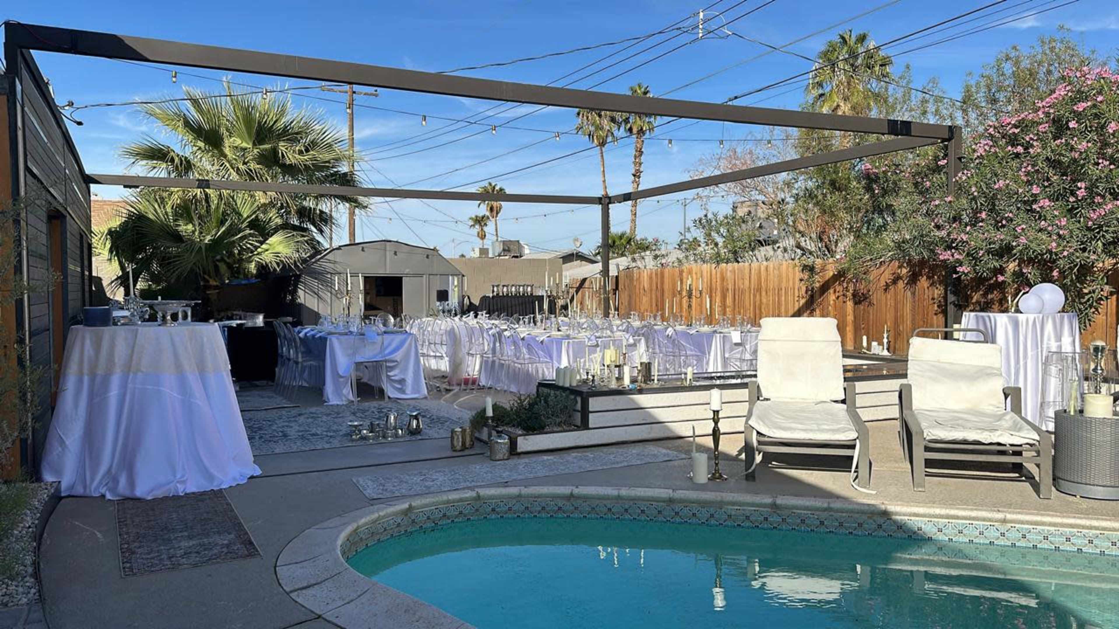 The image shows an outdoor venue set up for a celebration, featuring tables with white tablecloths, a pool, and a string of lights overhead.