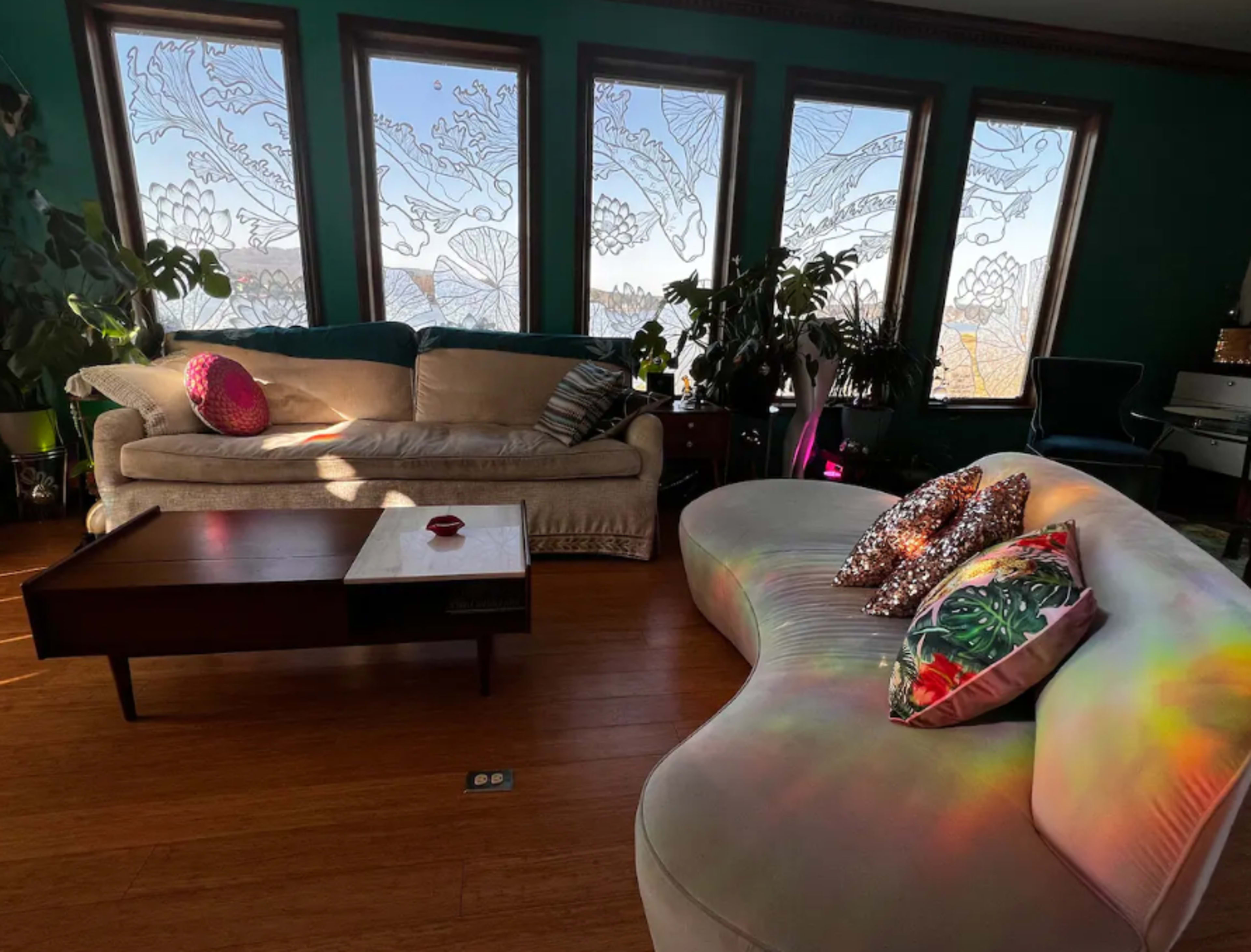 The image shows a living room with two distinct sofas, large windows adorned with intricate designs, and various plants scattered throughout the space.
