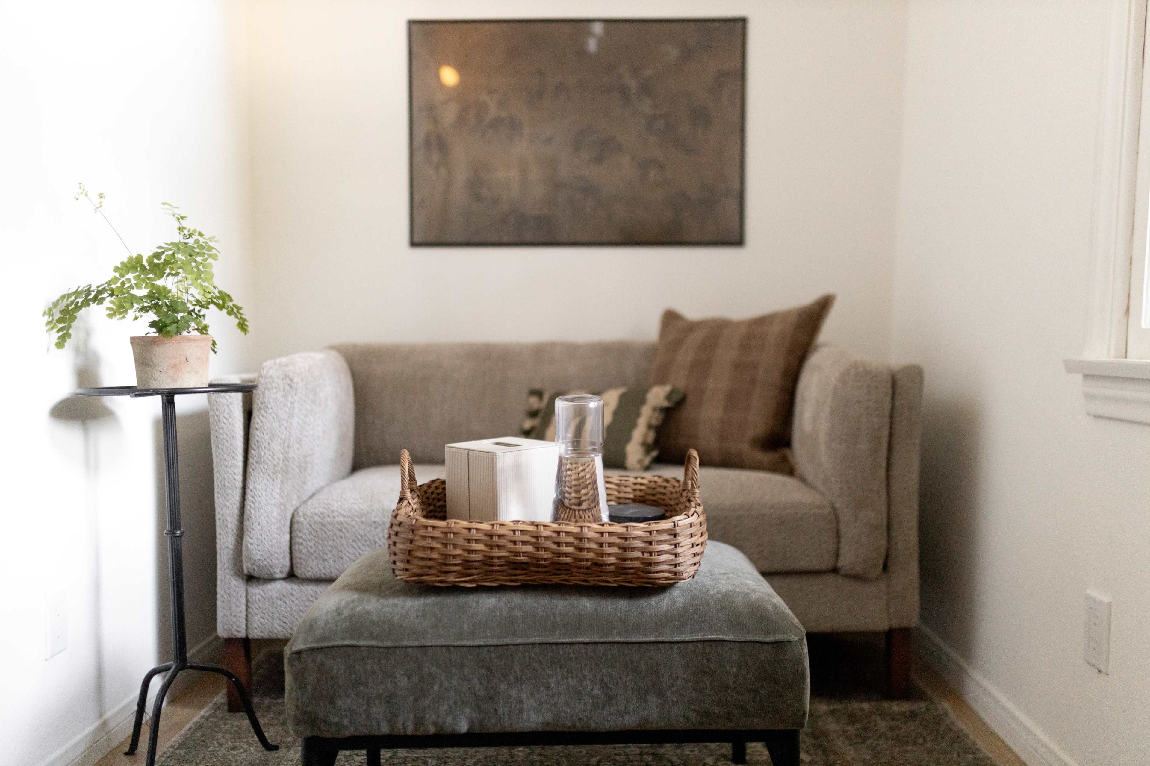 A cozy living space features a grey couch with decorative pillows, a small table with a potted plant, and a wicker basket on an ottoman, all set against a neutral wall.