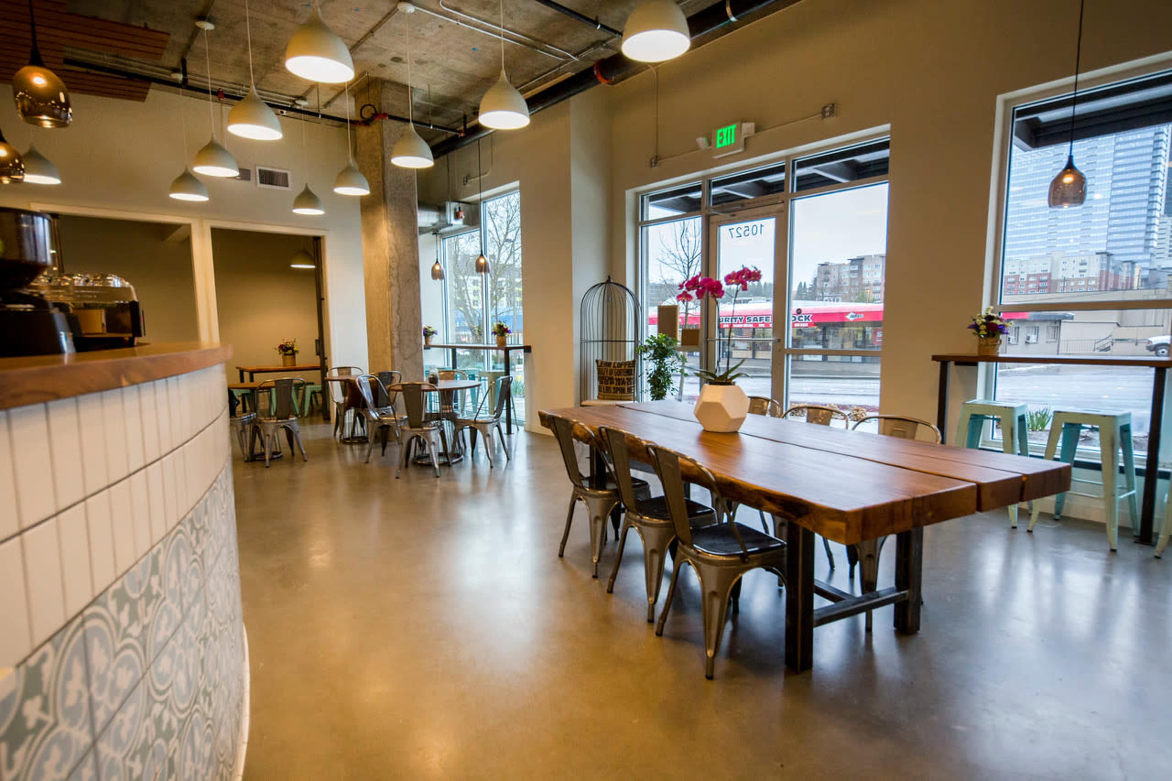 A modern café interior with a long wooden table, metal chairs, and large windows offering a view of the street outside.