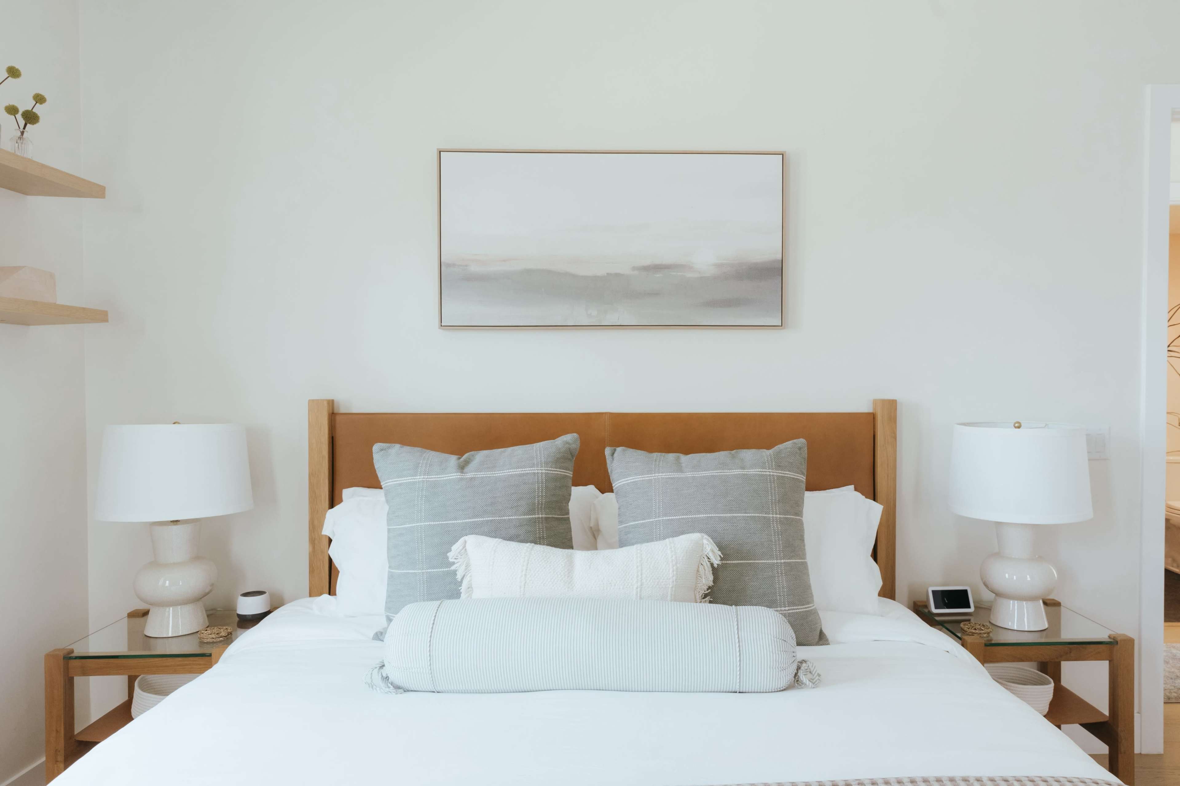 A neatly arranged bedroom features a wooden headboard with several gray and white pillows on the bed, flanked by two lamps on side tables.