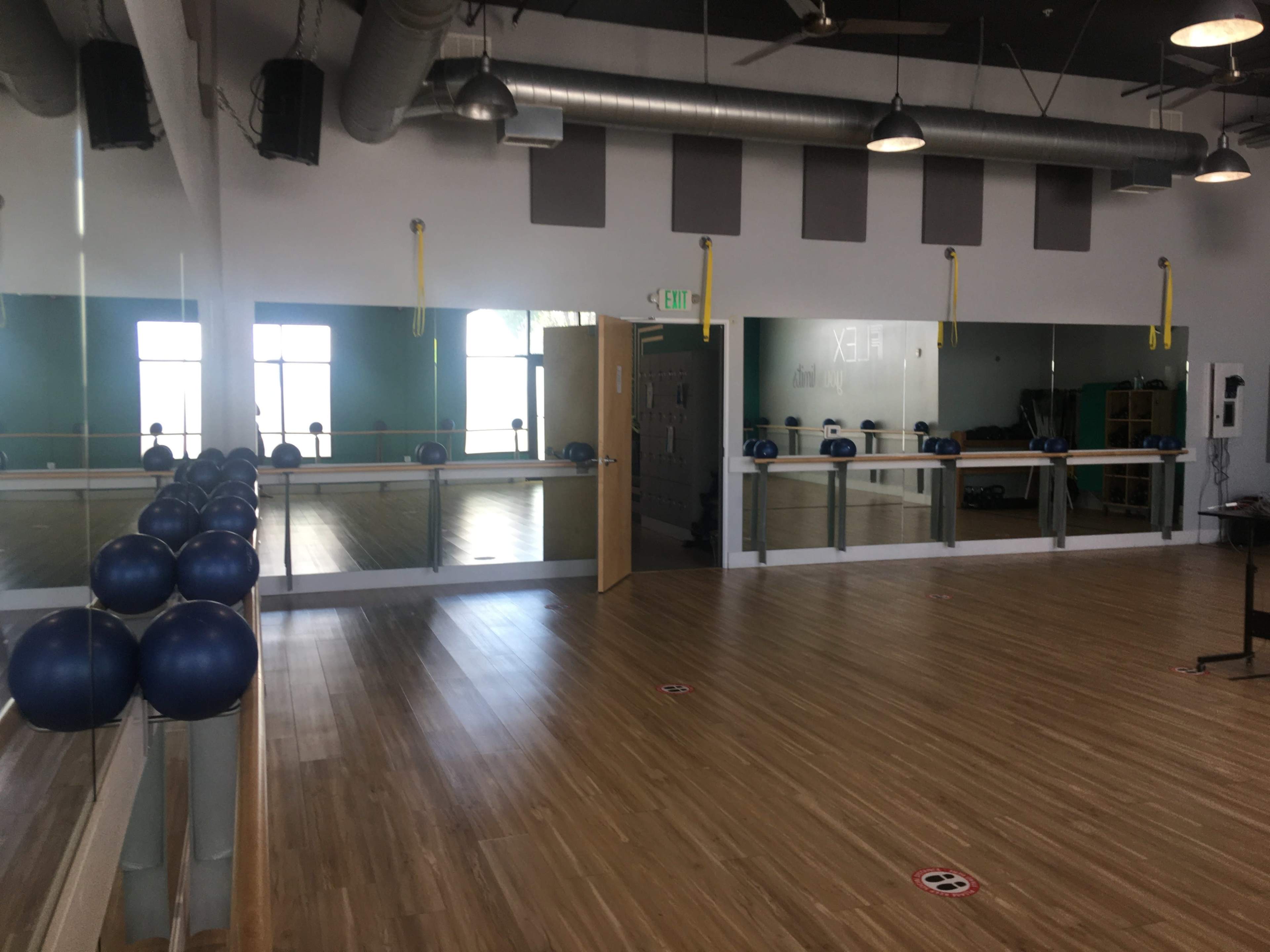 A spacious fitness studio features a mirrored wall, wooden flooring, exercise balls lined up along the bar, and marked social distancing circles on the floor.