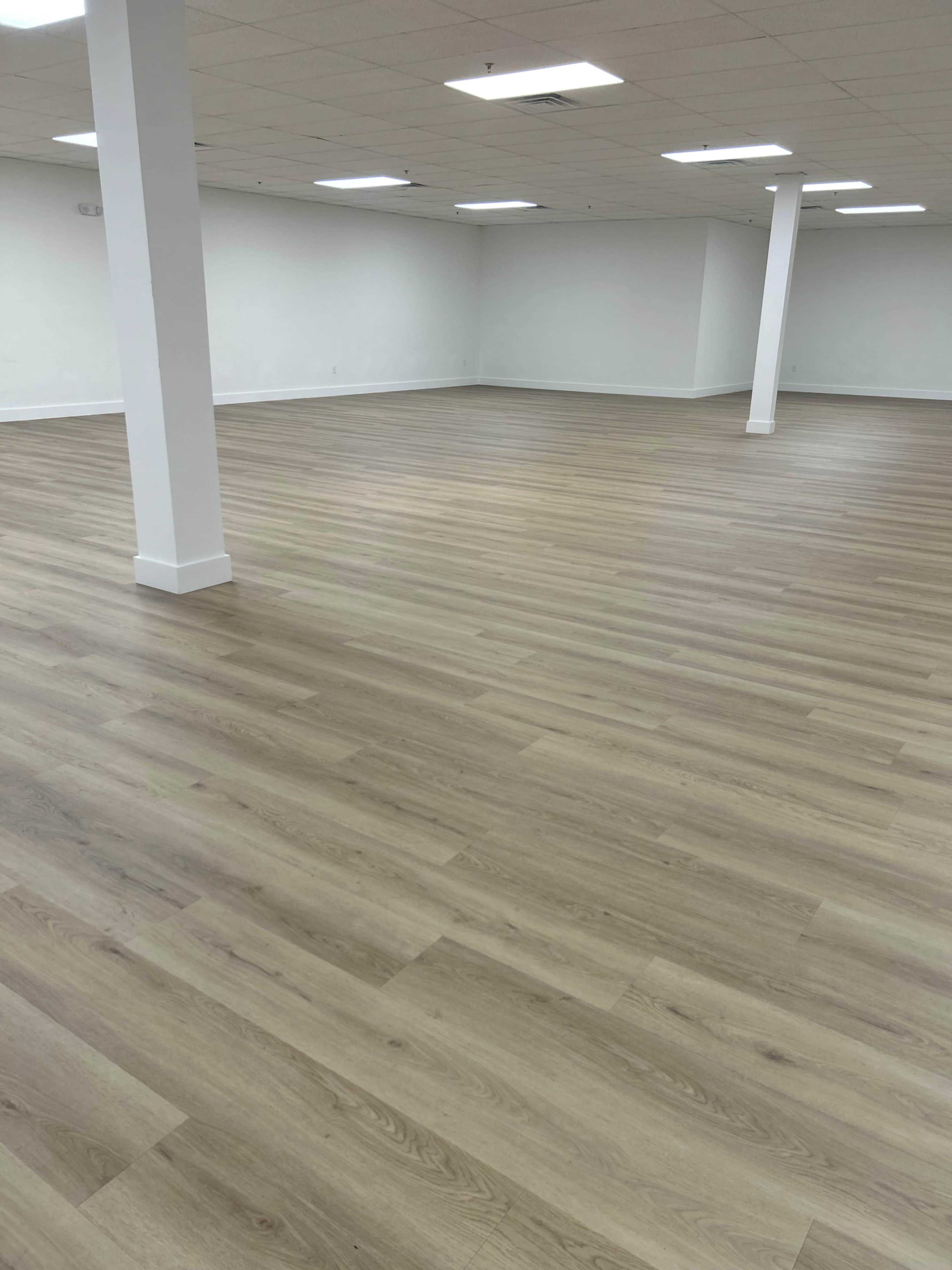 The image shows a spacious, empty room with light wood flooring and white walls, illuminated by ceiling lights.
