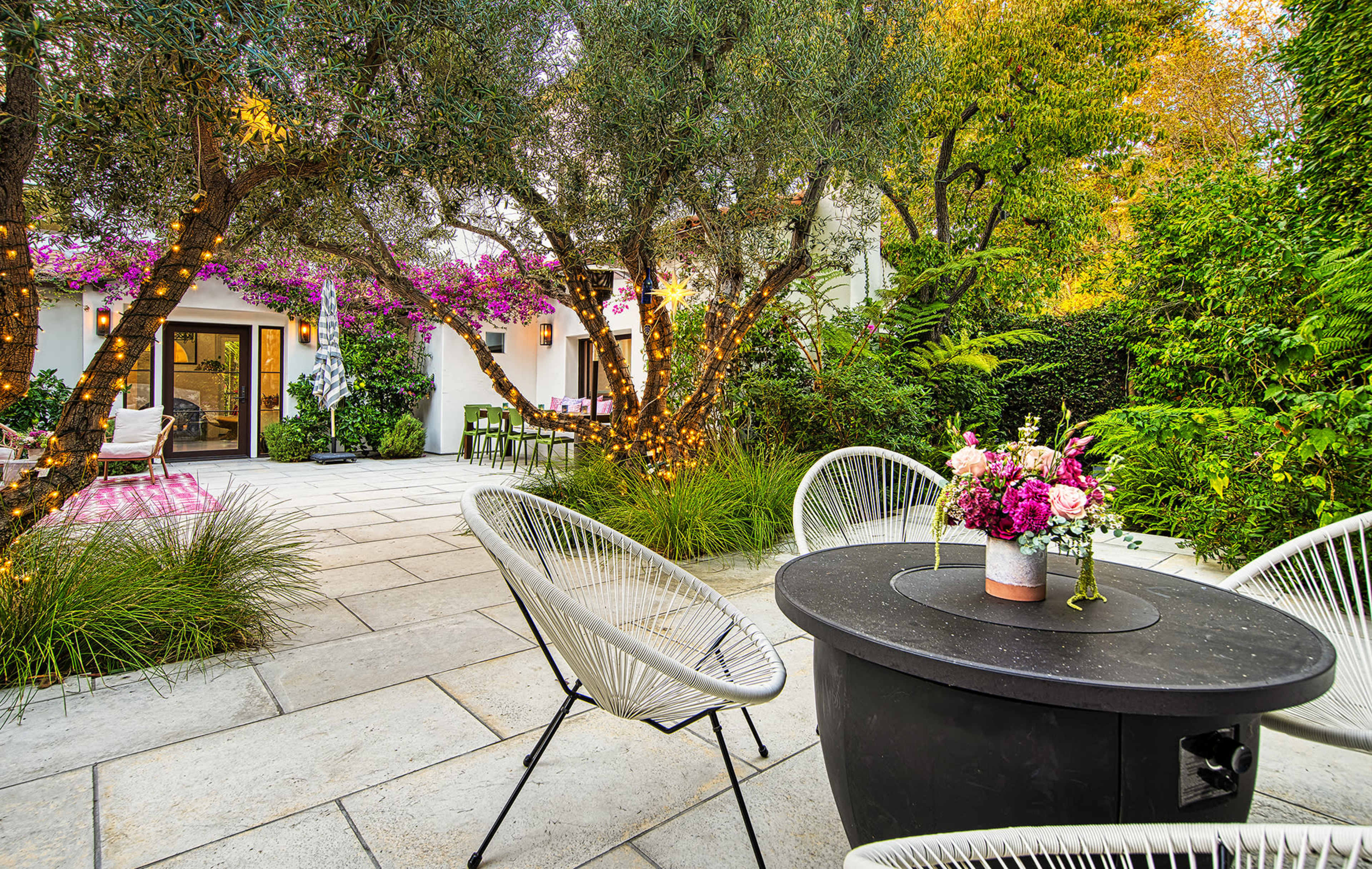 A landscaped outdoor seating area features a round black table surrounded by white chairs, with flowering plants and olive trees in the background.