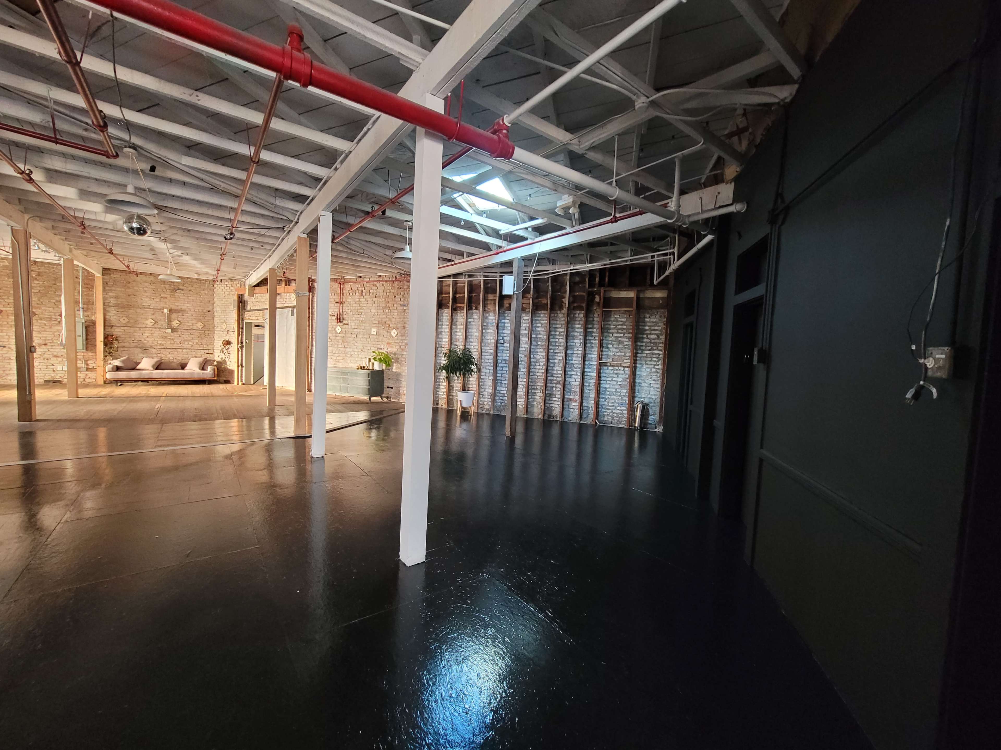 The image shows an open, spacious interior of a studio with brick walls, polished black floors, and exposed beams.