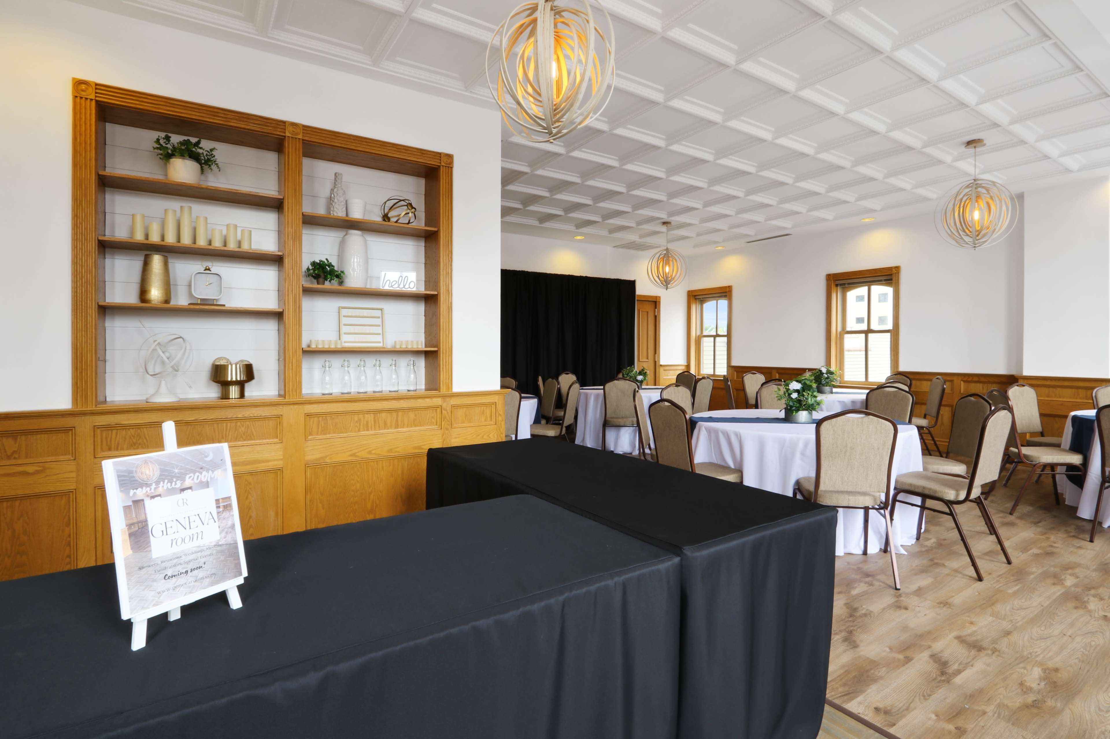 A well-lit event space featuring tables with beige chairs, a black table in the foreground, and a decorative shelving unit against the wall.