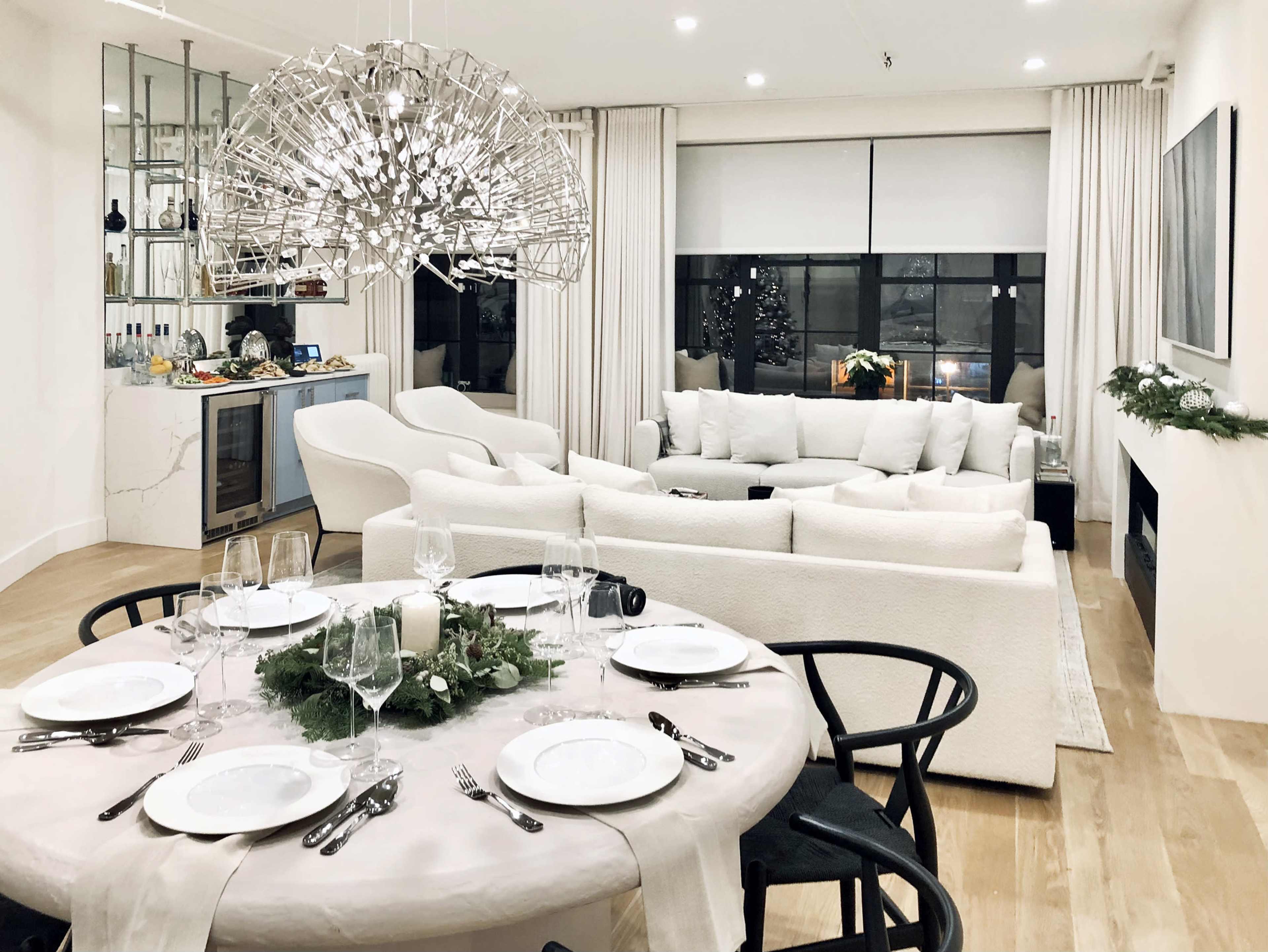 A dining table is set with plates and glassware in the foreground, while a modern living room with a large chandelier and a cozy seating area is visible in the background.