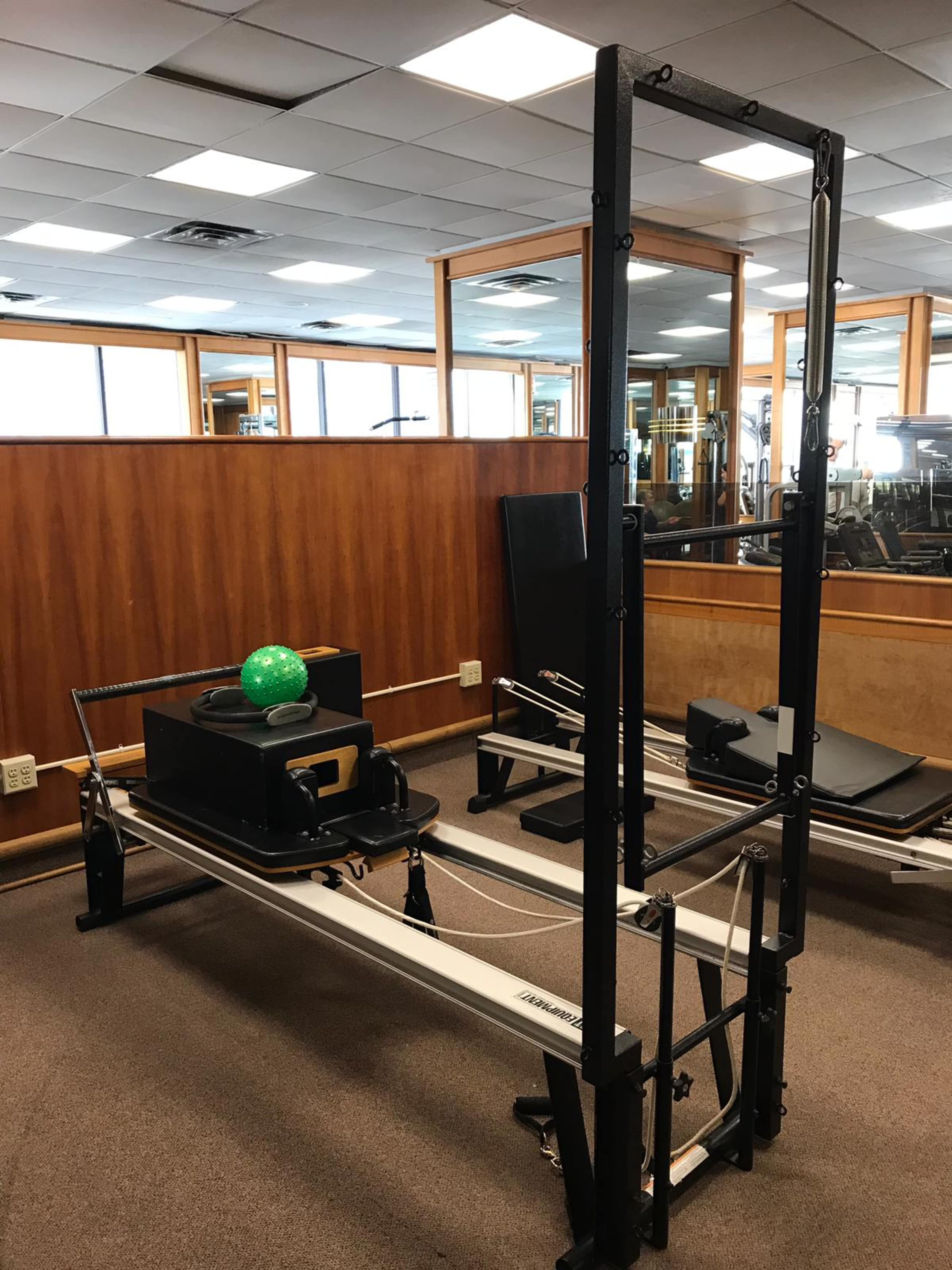 A Pilates reformer machine with a green stability ball placed on top, situated in a gym with wooden paneling and mirrors in the background.