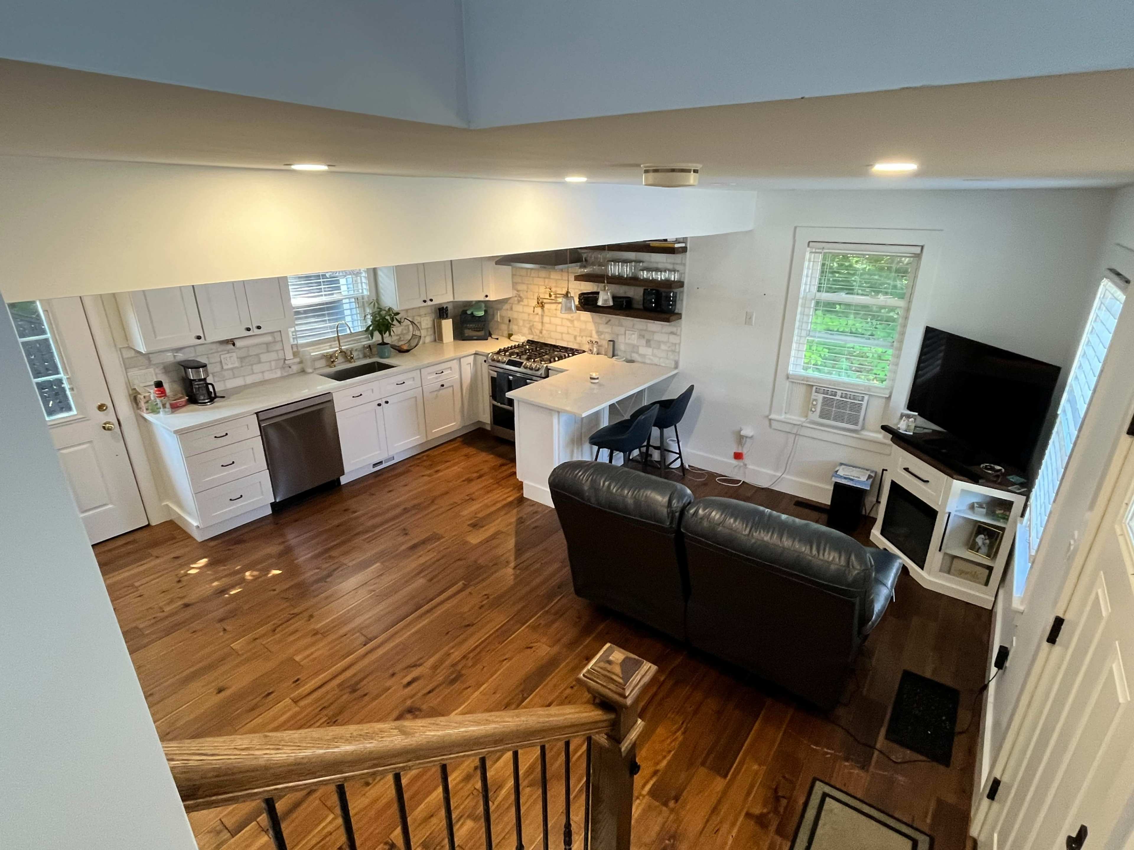 The image shows a modern kitchen and living area featuring wooden floors, white cabinetry, and a seating area with a couch and an entertainment unit.