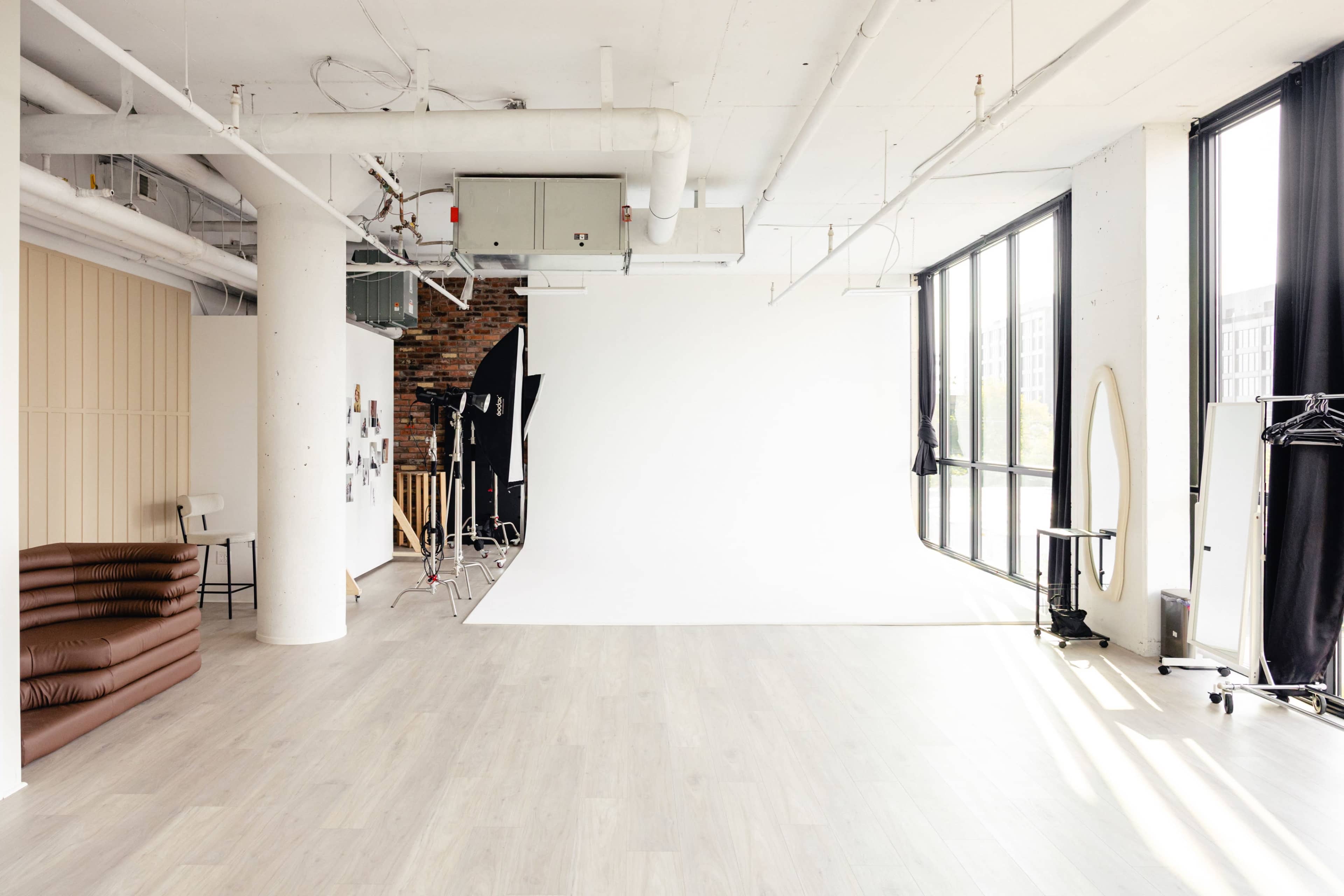 The image shows a spacious studio with large windows, featuring a blank white backdrop, wooden flooring, and various studio equipment.