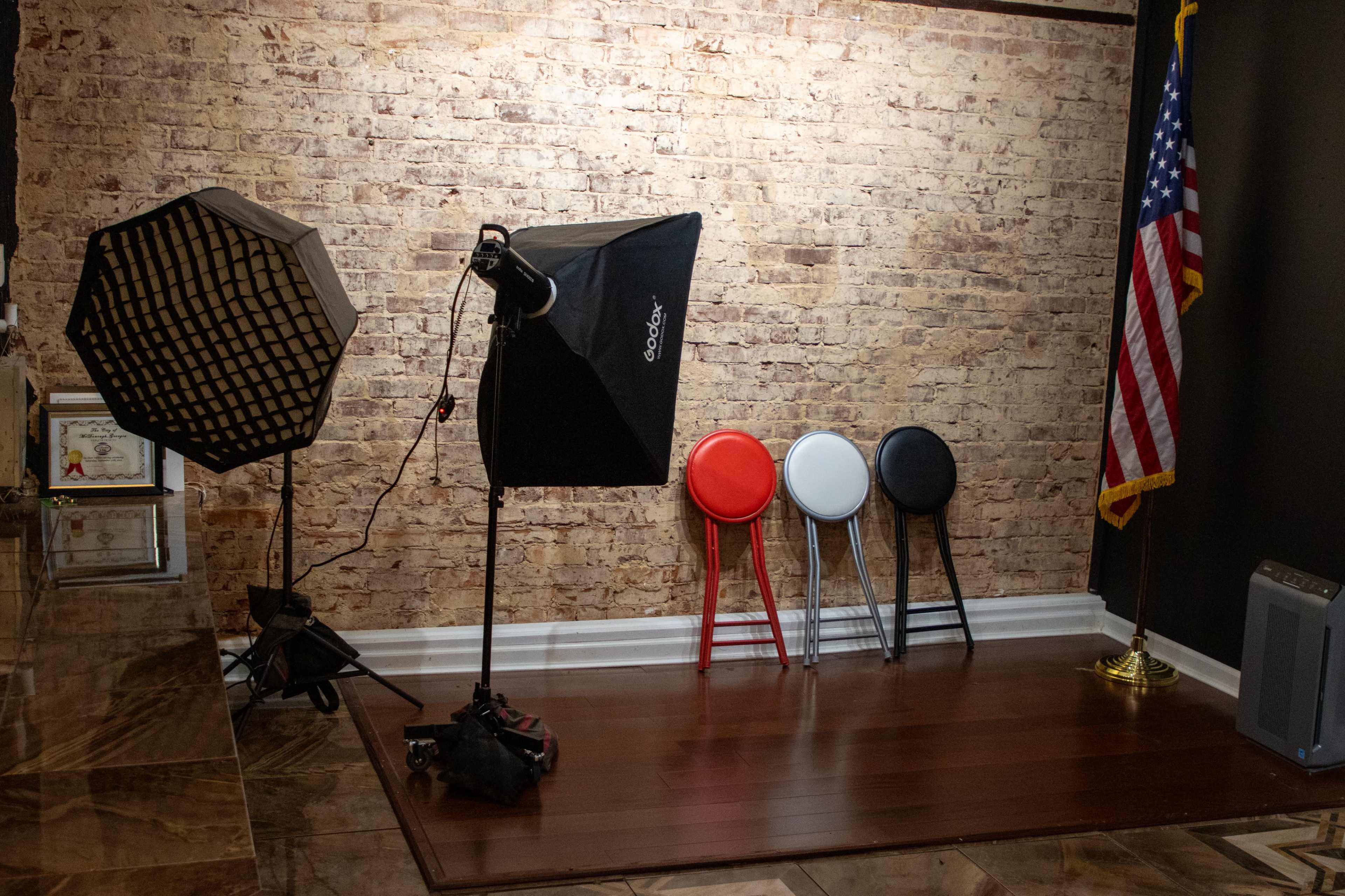 A photography setup with light fixtures, three stools in red, white, and black, and an American flag against a brick wall.