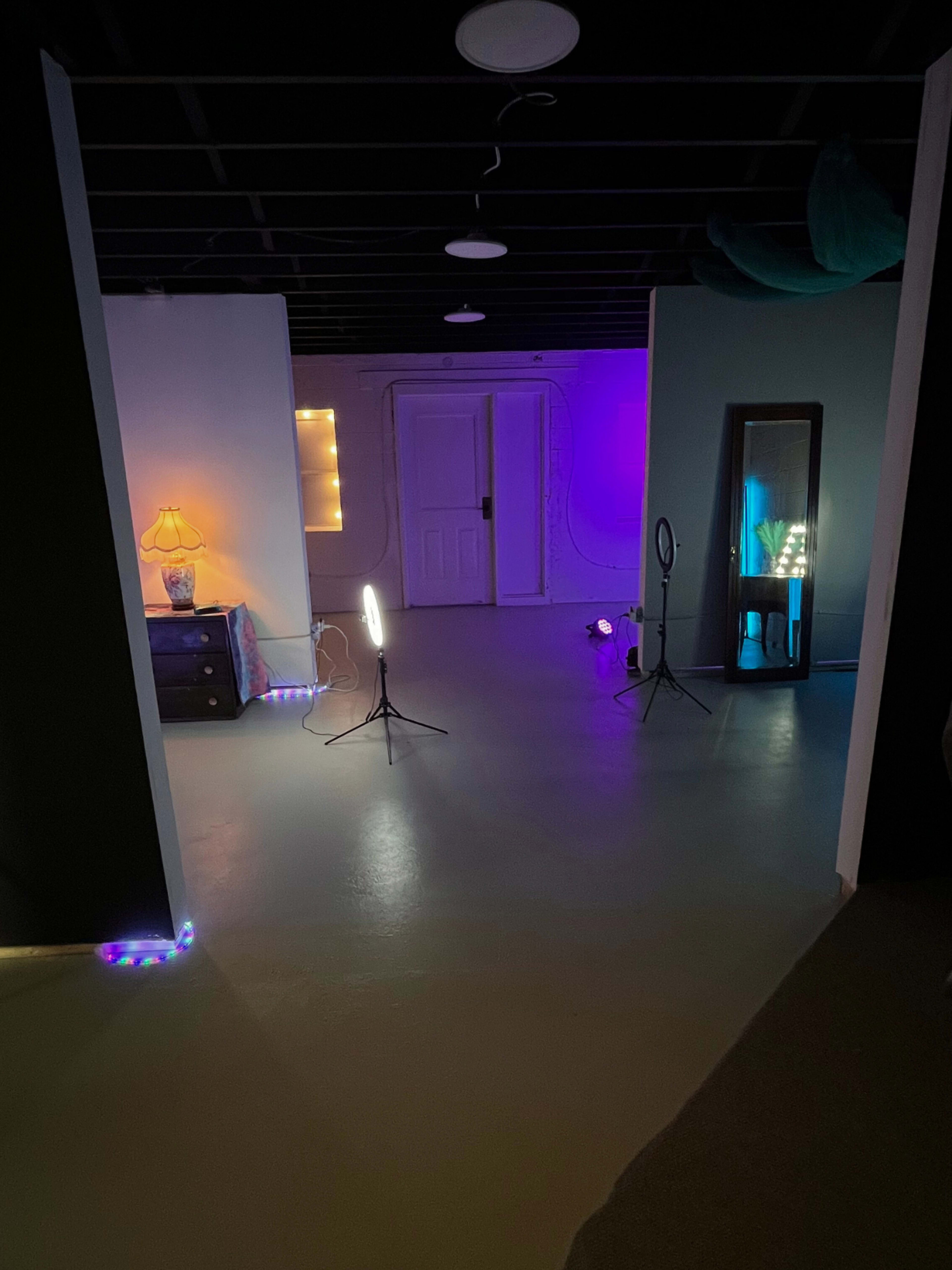 A dimly lit room with colorful lighting featuring a mirror, a lamp, and two light stands set up for photography or video use.