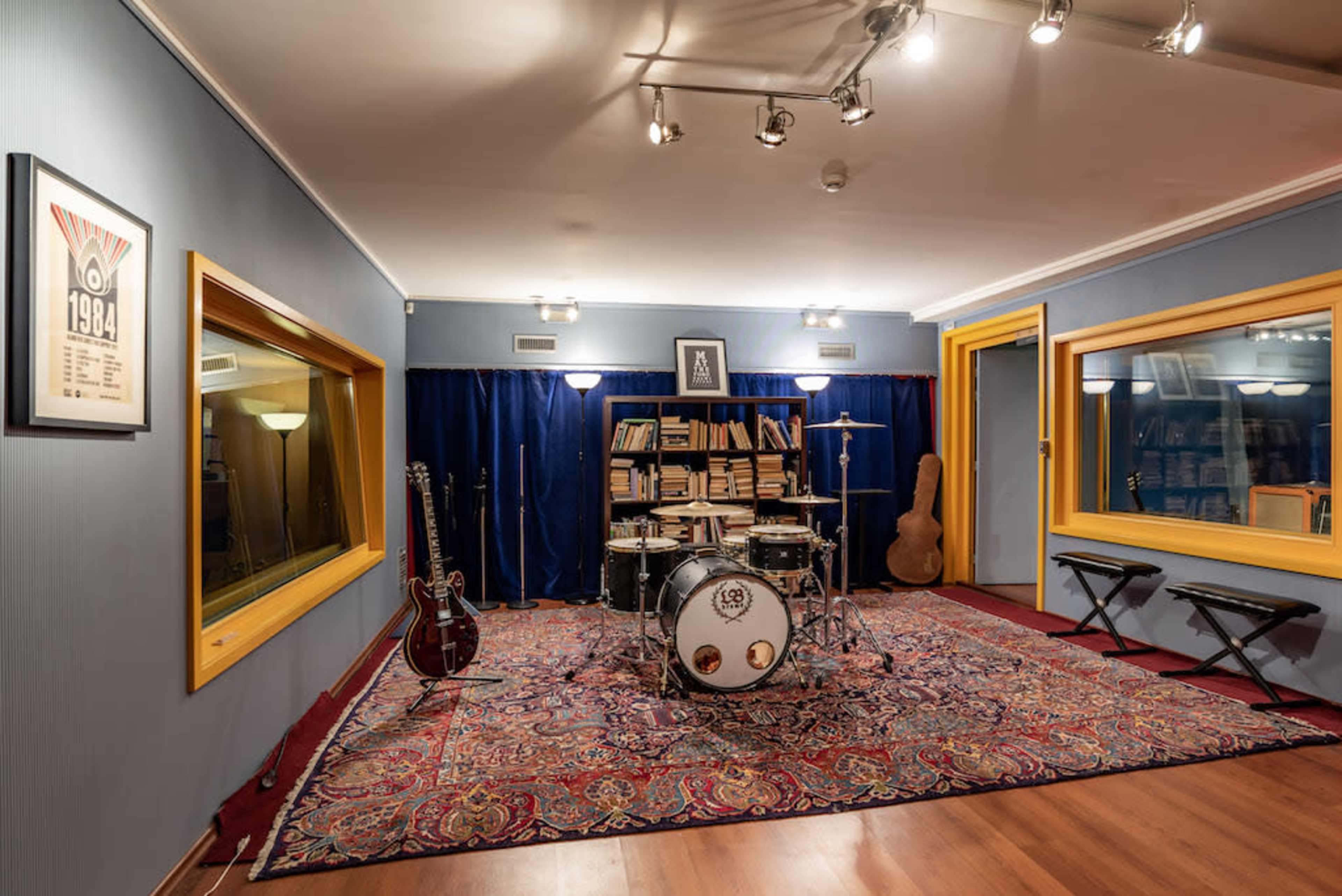 A music studio featuring a drum set, electric guitar, two keyboards, and shelves filled with books, all arranged on a decorative rug.