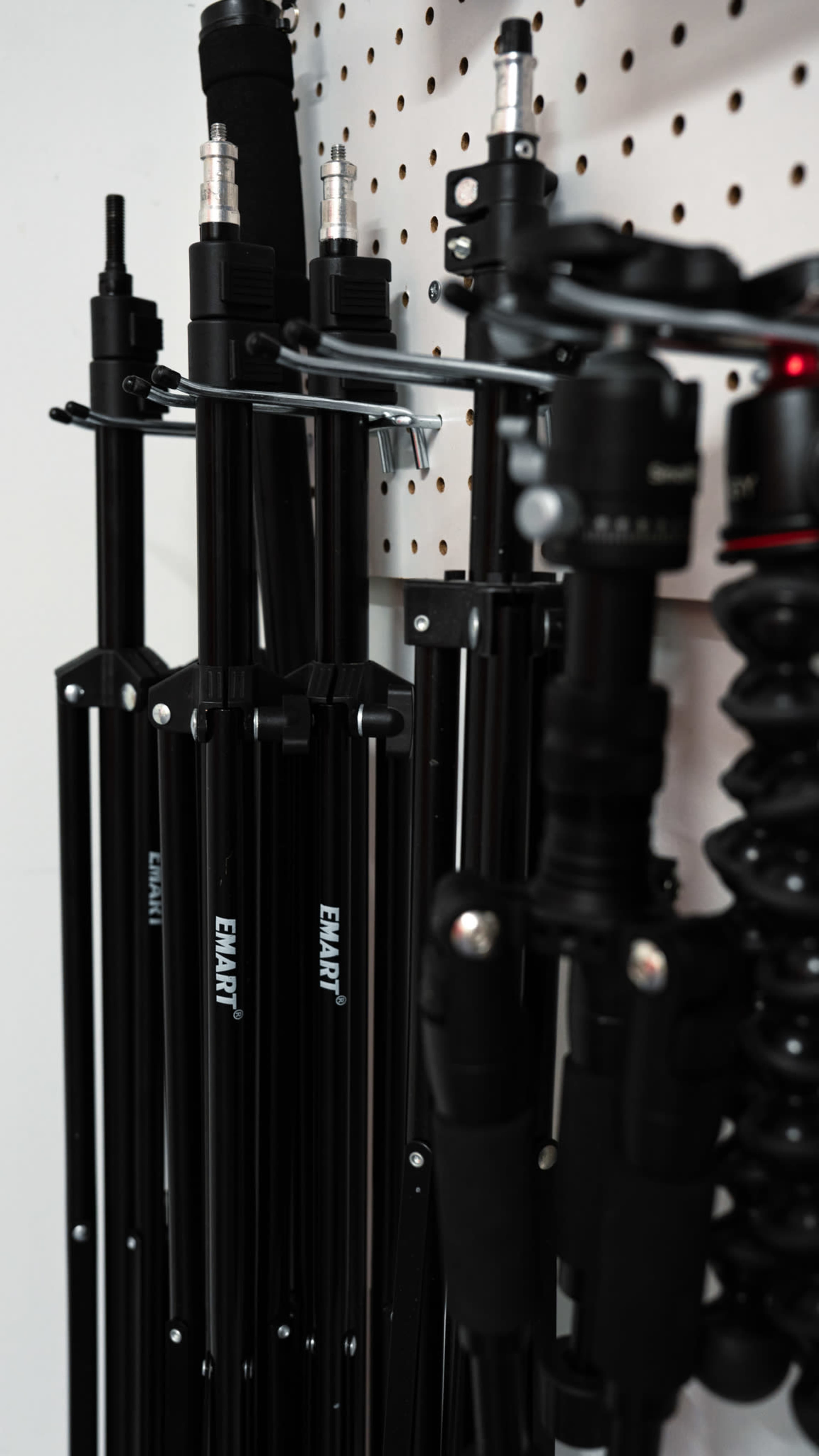 A collection of black photography tripods and support equipment is mounted on a pegboard wall.