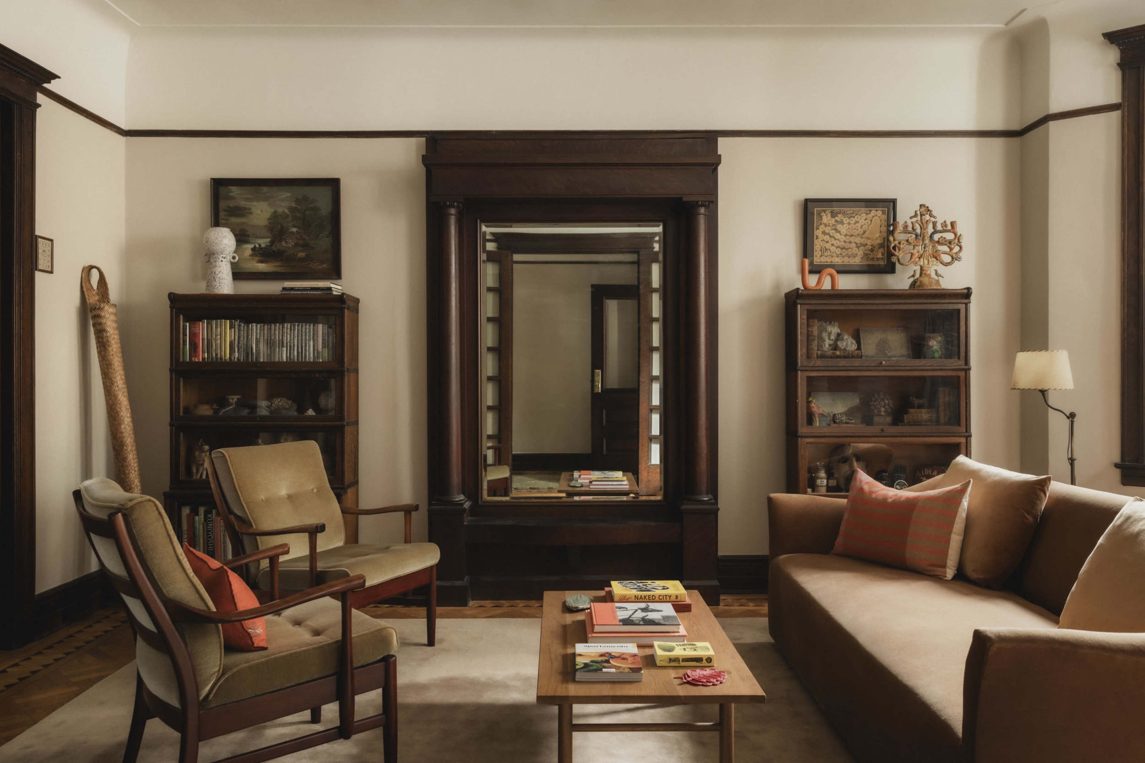 The image shows a cozy living room featuring a brown sofa, wooden bookshelves, a mirror, and a coffee table with various items on it.