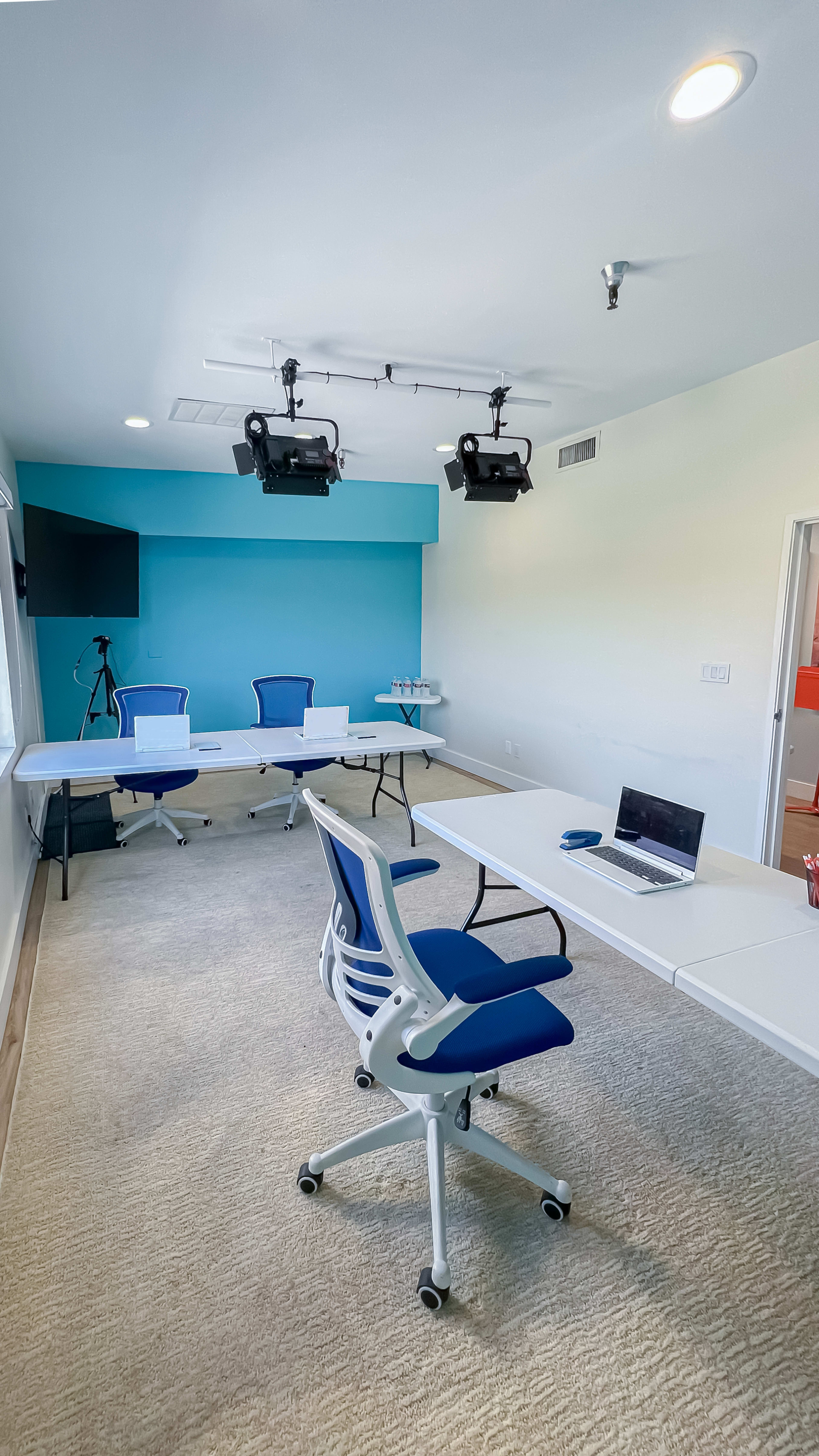 The image features a modern conference room with white tables, blue chairs, a laptop on one table, and projectors mounted on the ceiling.
