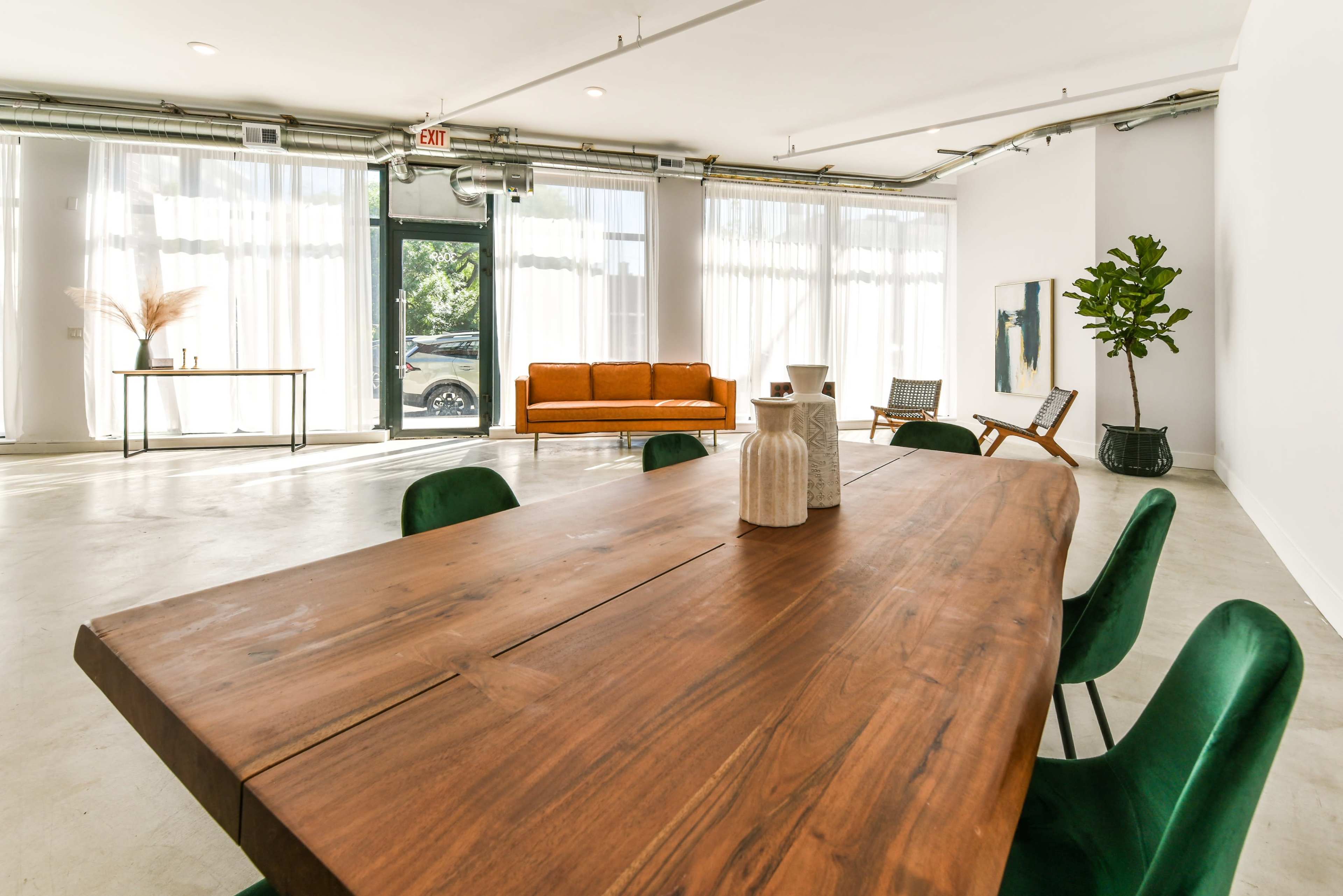 A spacious, well-lit room features a large wooden dining table surrounded by green chairs, with a sofa, artwork, and a potted plant in the background.