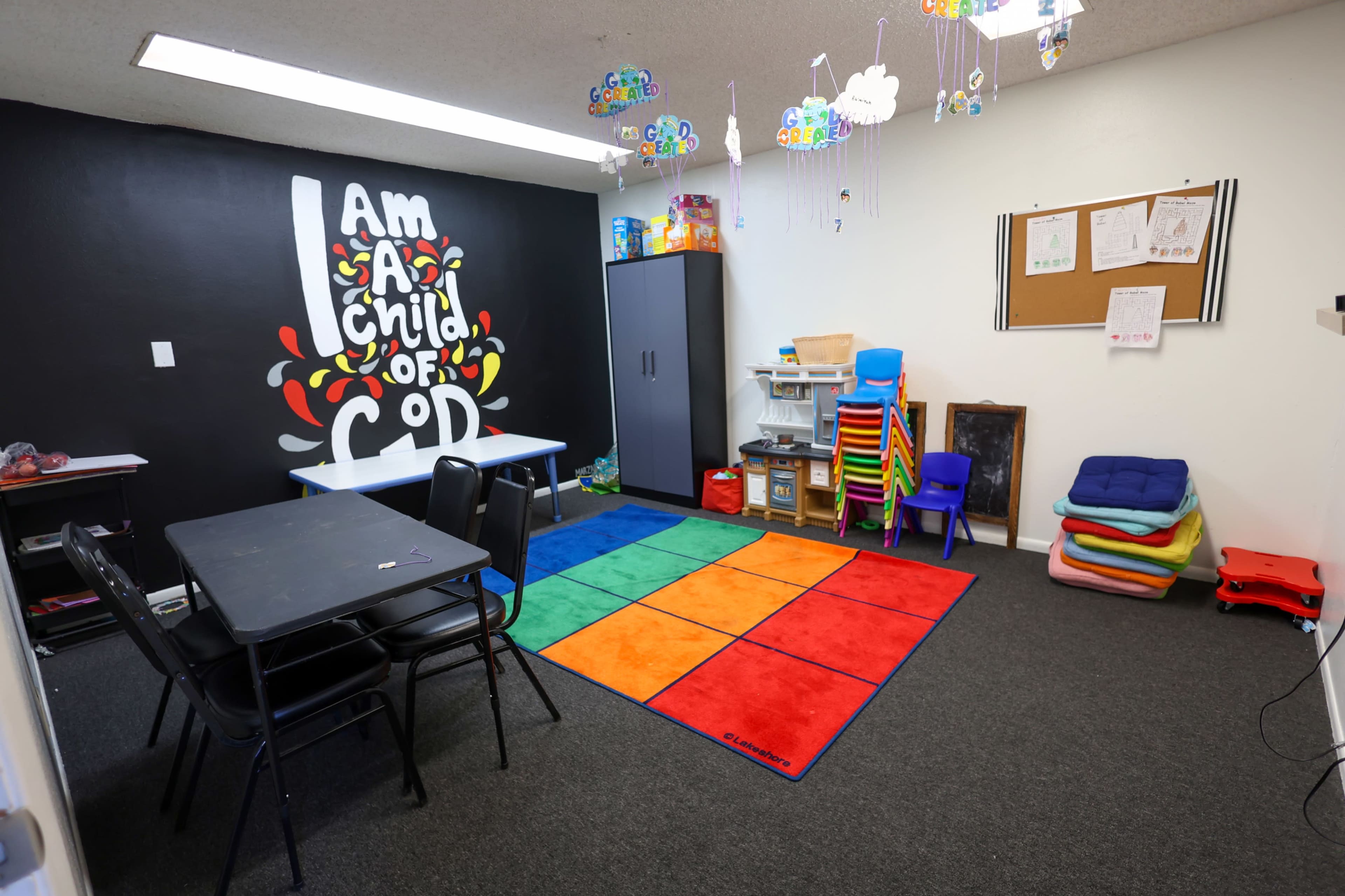 The image shows a children's classroom featuring a colorful carpet, desks, storage units, and a black wall with the text "I Am A Child of God."