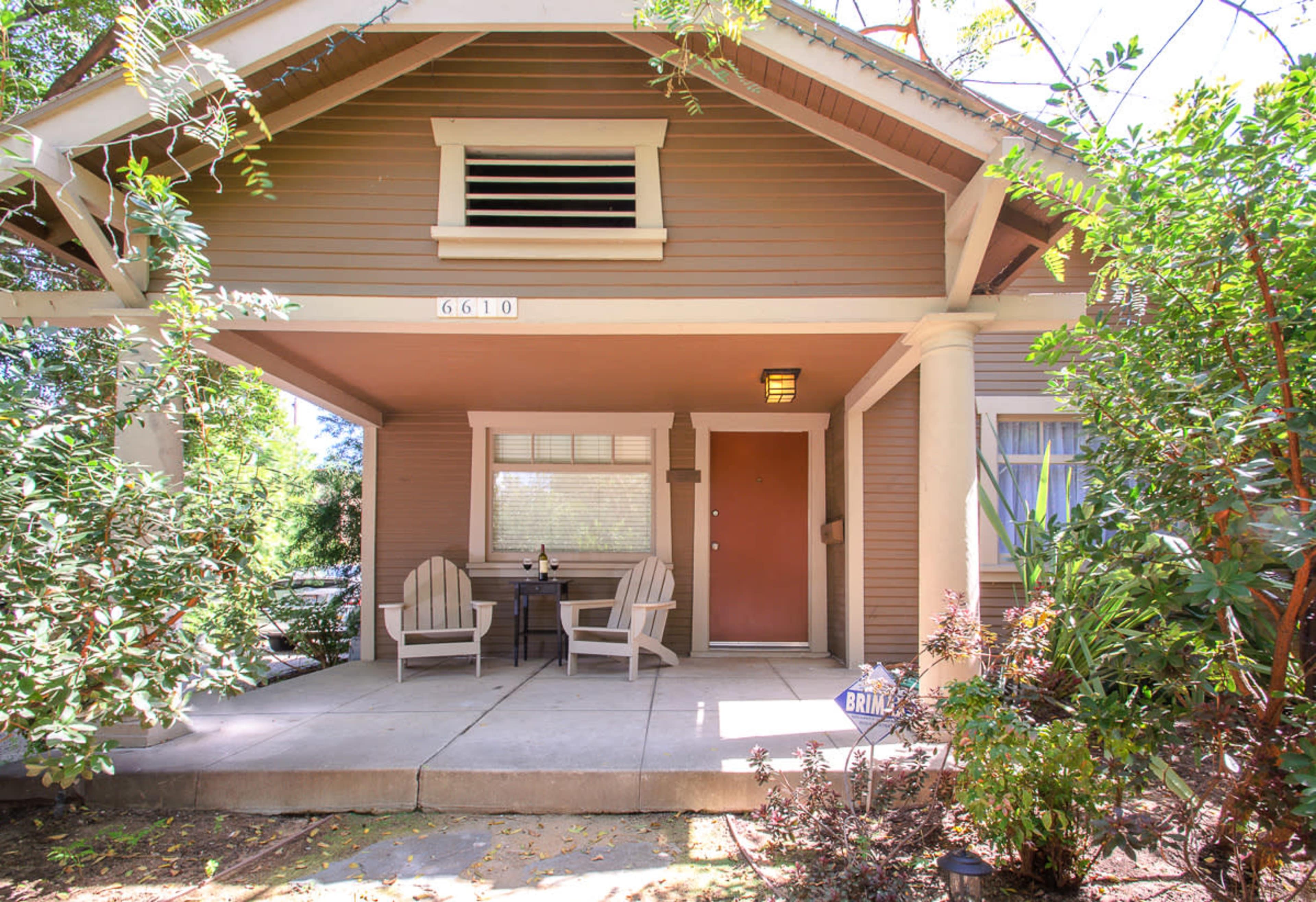 A front porch of a house with two chairs and a small table, surrounded by greenery.