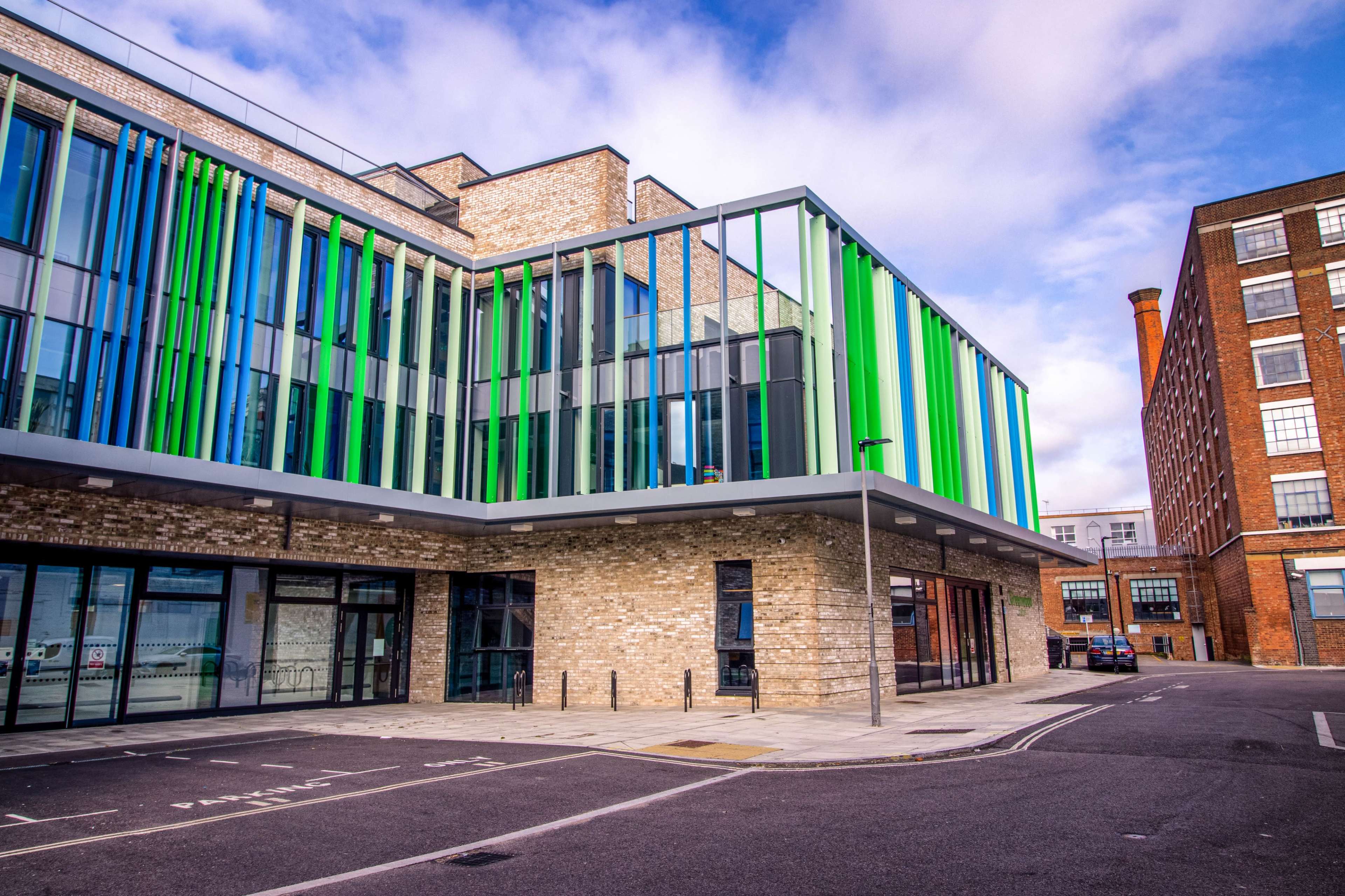 A modern building with colorful vertical glass panels and a brick facade, situated on a street next to a brick factory.
