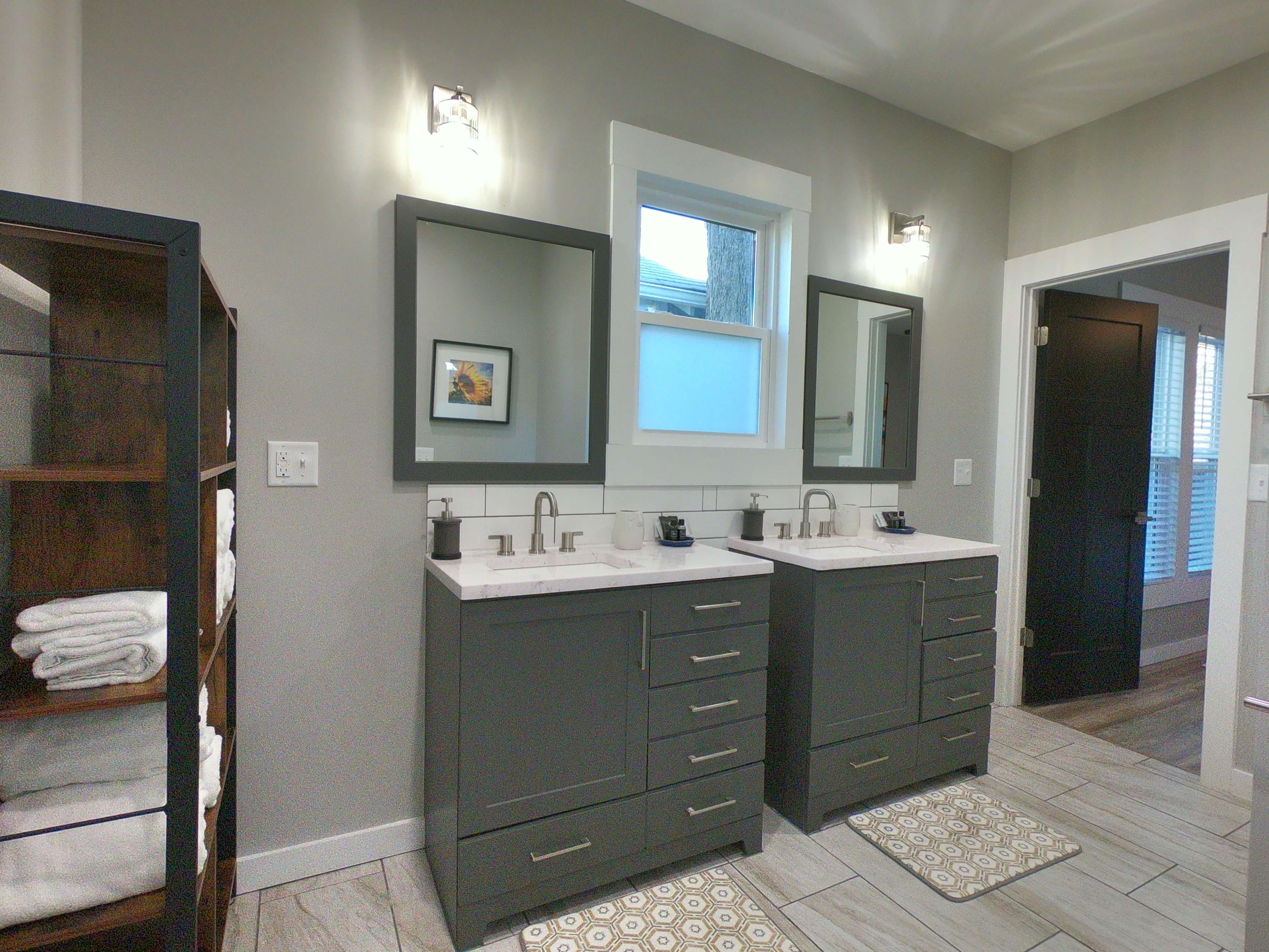 A modern bathroom featuring dual sinks with dark cabinetry, light-colored countertops, and a tall shelving unit with neatly stacked towels.
