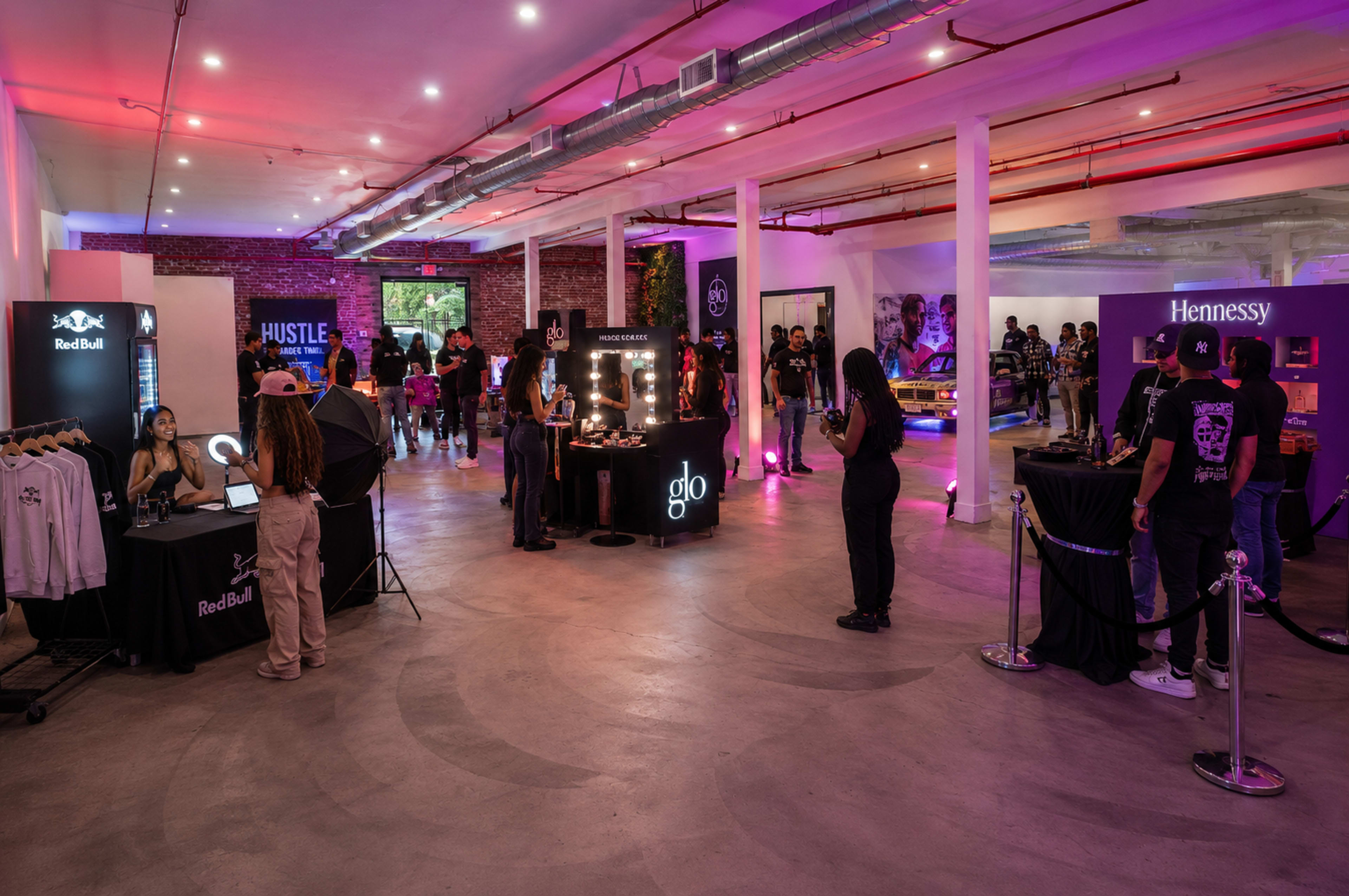 The image shows an indoor event space with various booths for brands like Red Bull and Hennessy, where guests interact and explore the setups amidst ambient lighting.