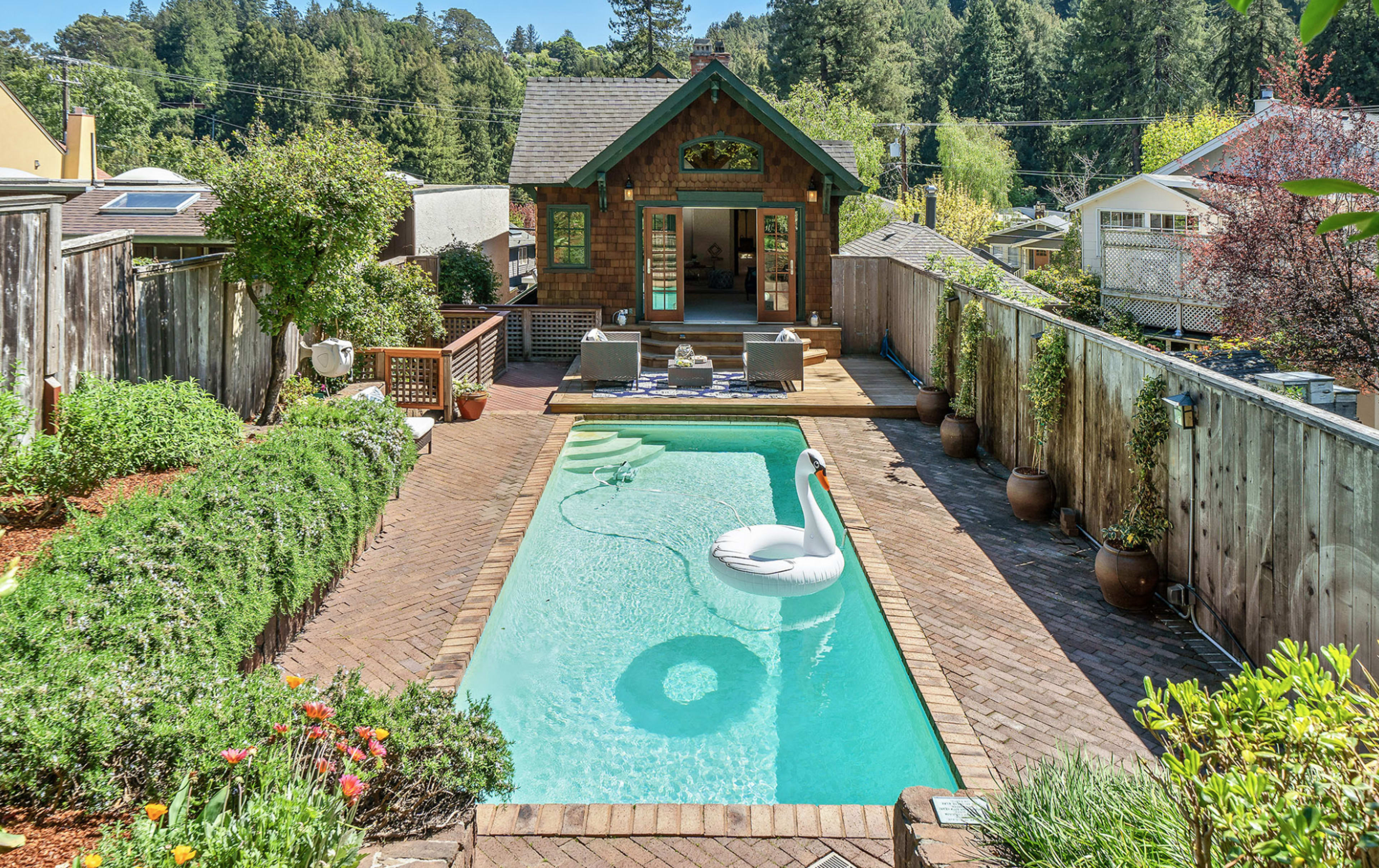 A backyard features a rectangular pool with a swan float, surrounded by lush greenery and leading to a small house with large windows.