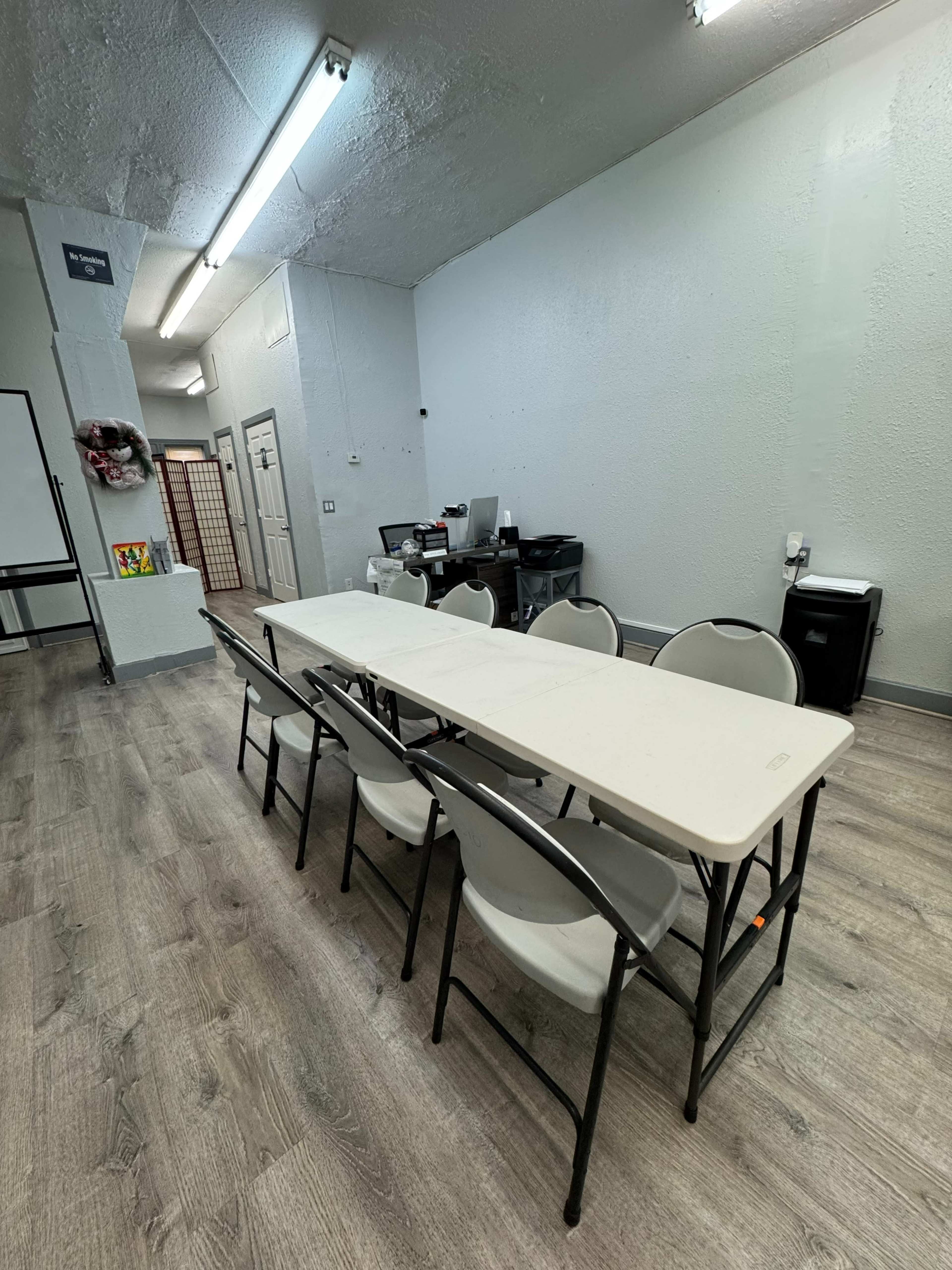 The image shows a spacious room with a long folding table surrounded by chairs, and various office equipment along the back wall.