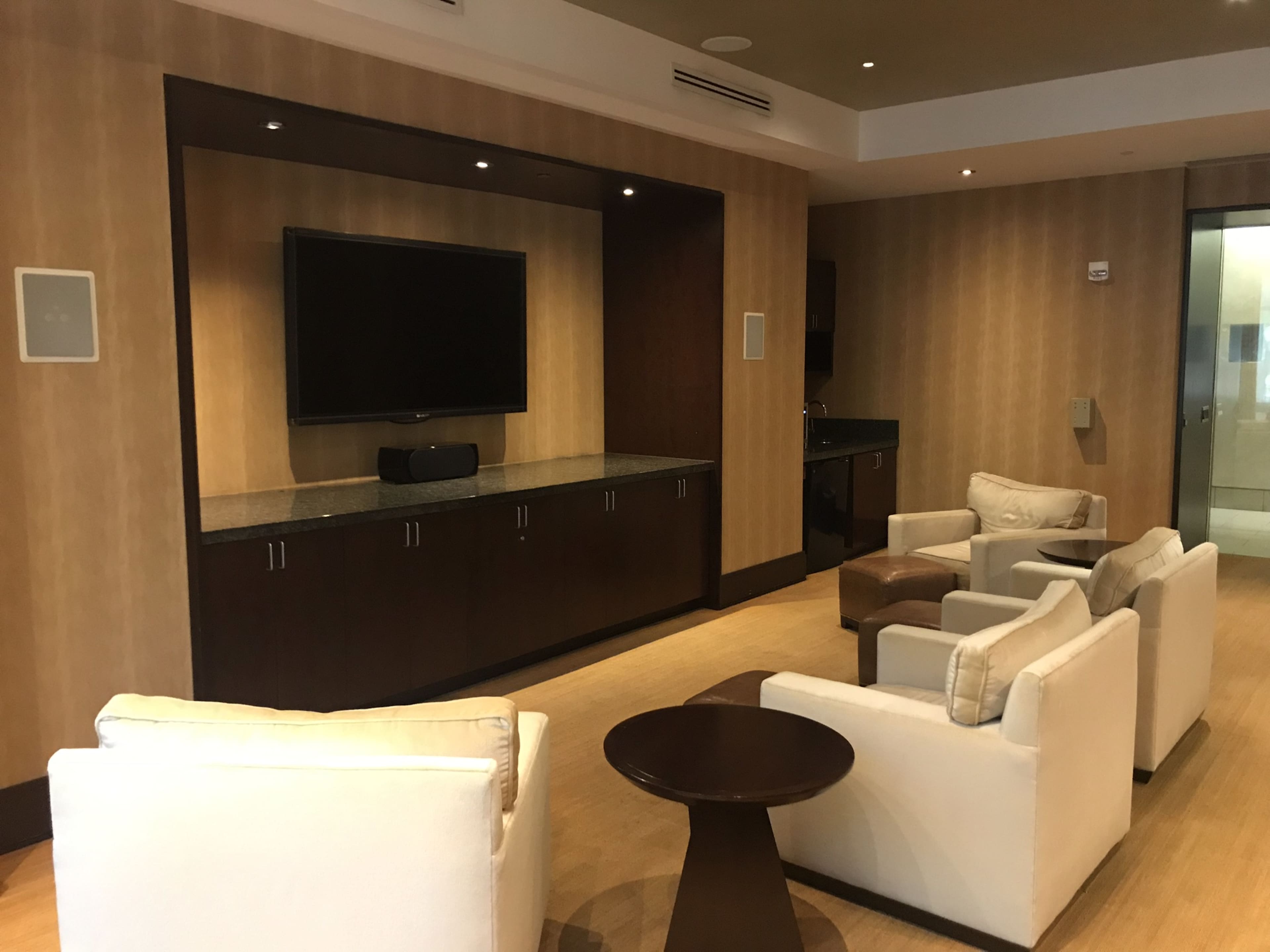 A modern lounge area with two armchairs and a sofa arranged around a low table, facing a wall-mounted television and a dark wood cabinet.