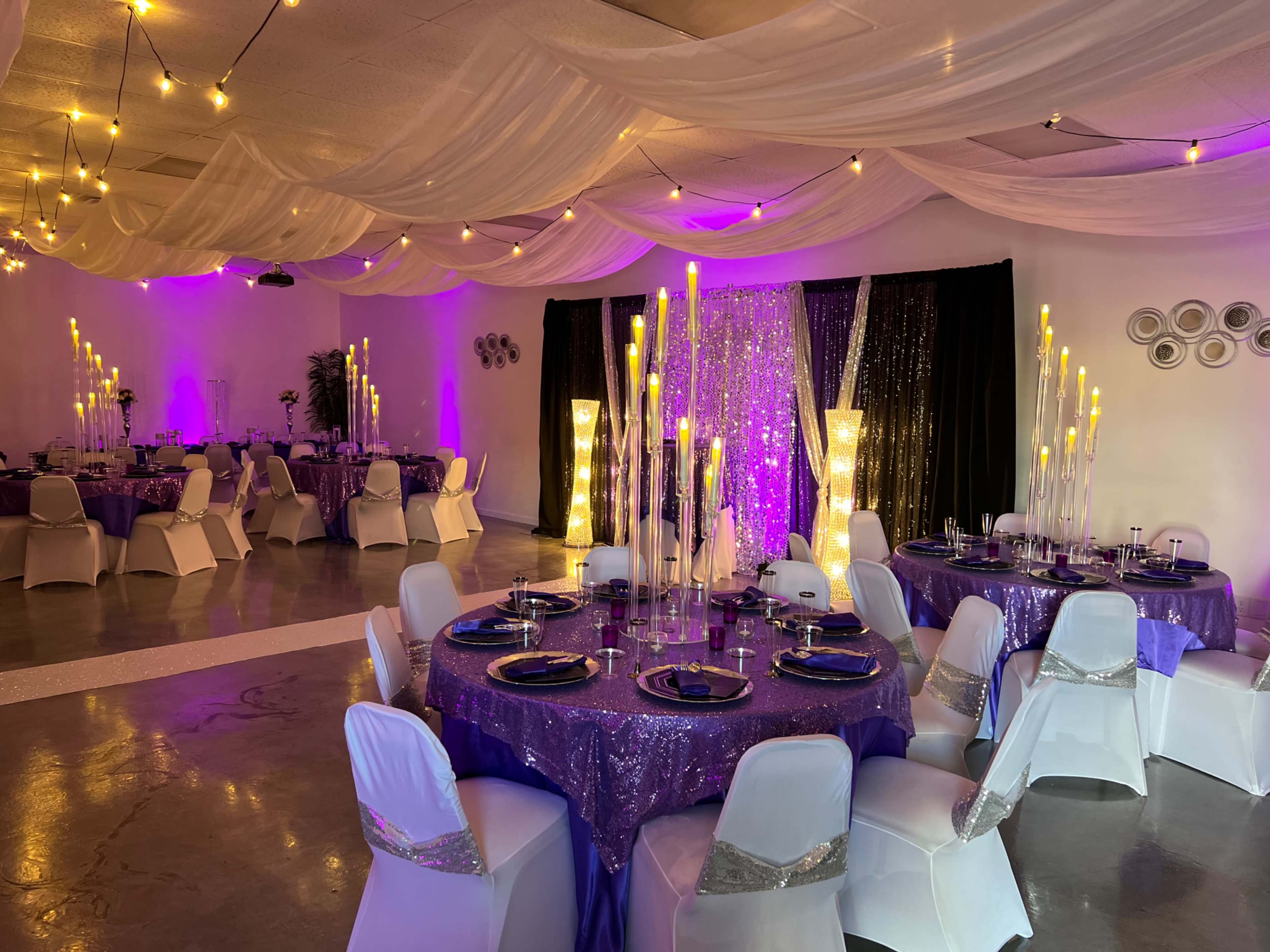 A decorated event space with tables covered in purple cloth, elegant candle centers, and ambient lighting from draped fabric and string lights.