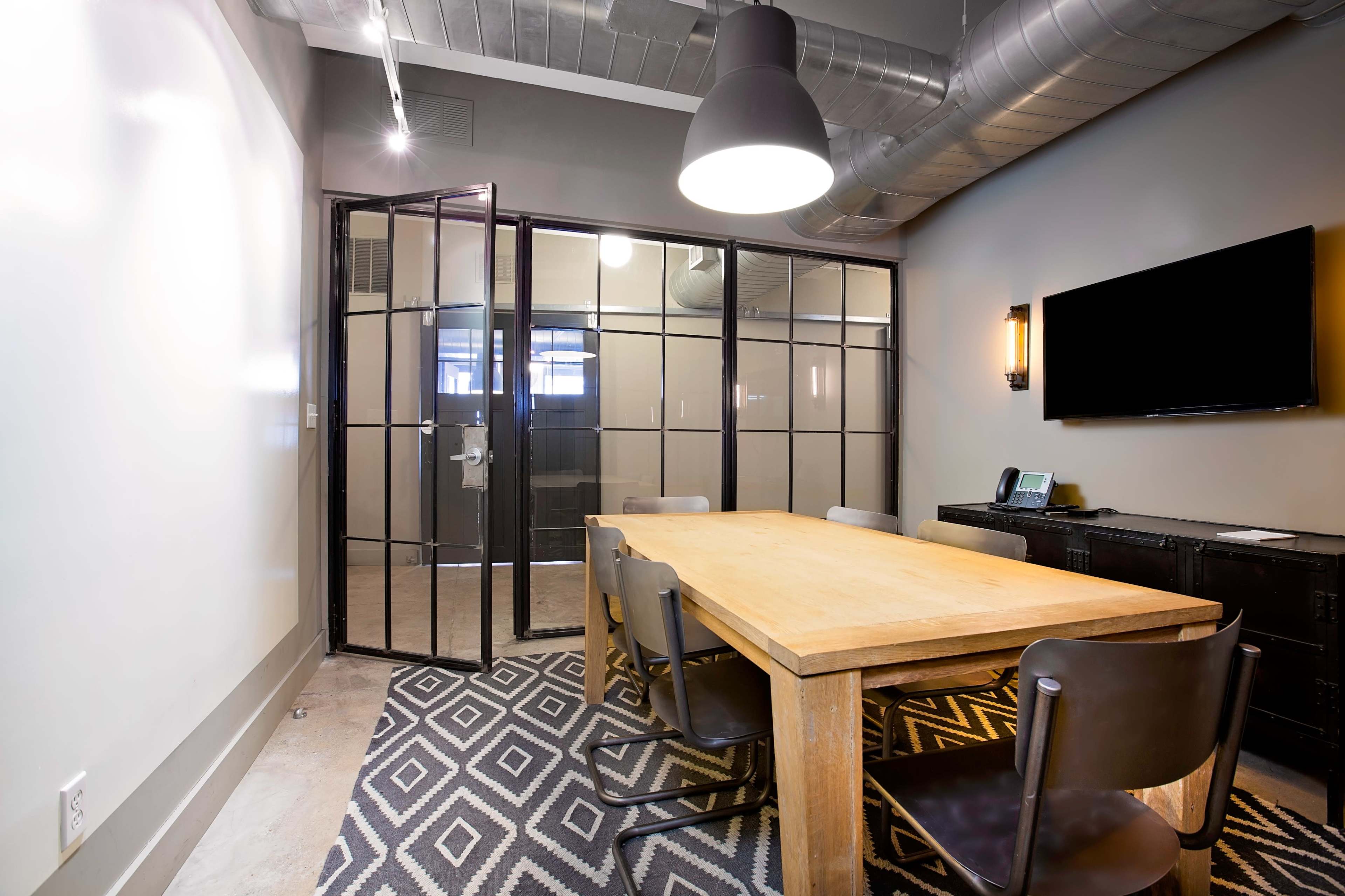 The image shows a modern conference room with a wooden table surrounded by chairs, glass walls, and a wall-mounted TV.