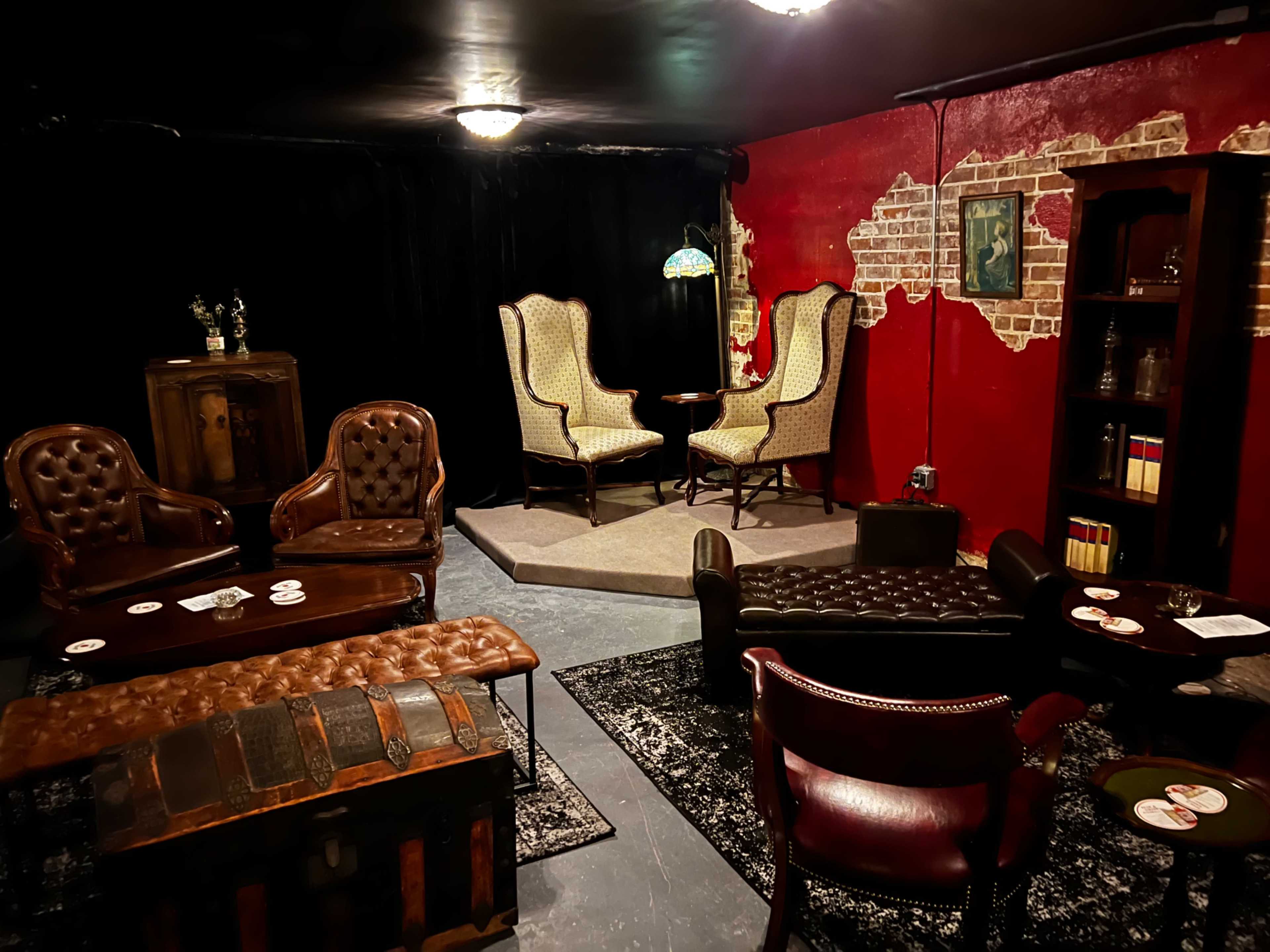 A dimly lit lounge area with dark walls, featuring leather armchairs, a coffee table, and a bookshelf against a red-brick wall.