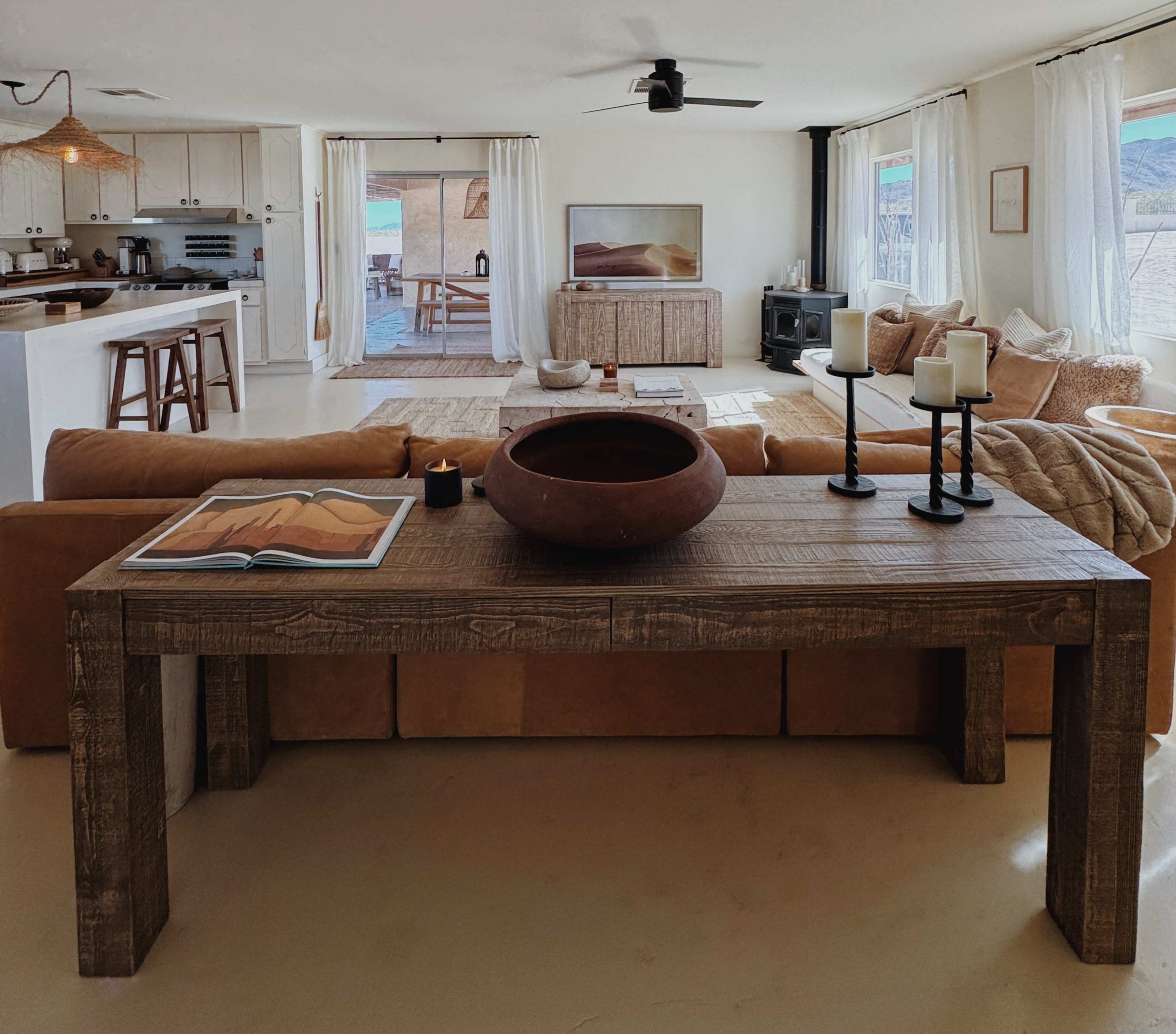 The image shows a spacious, modern living room featuring a wooden table with a bowl and candles, a large sofa, and large windows overlooking a patio area.