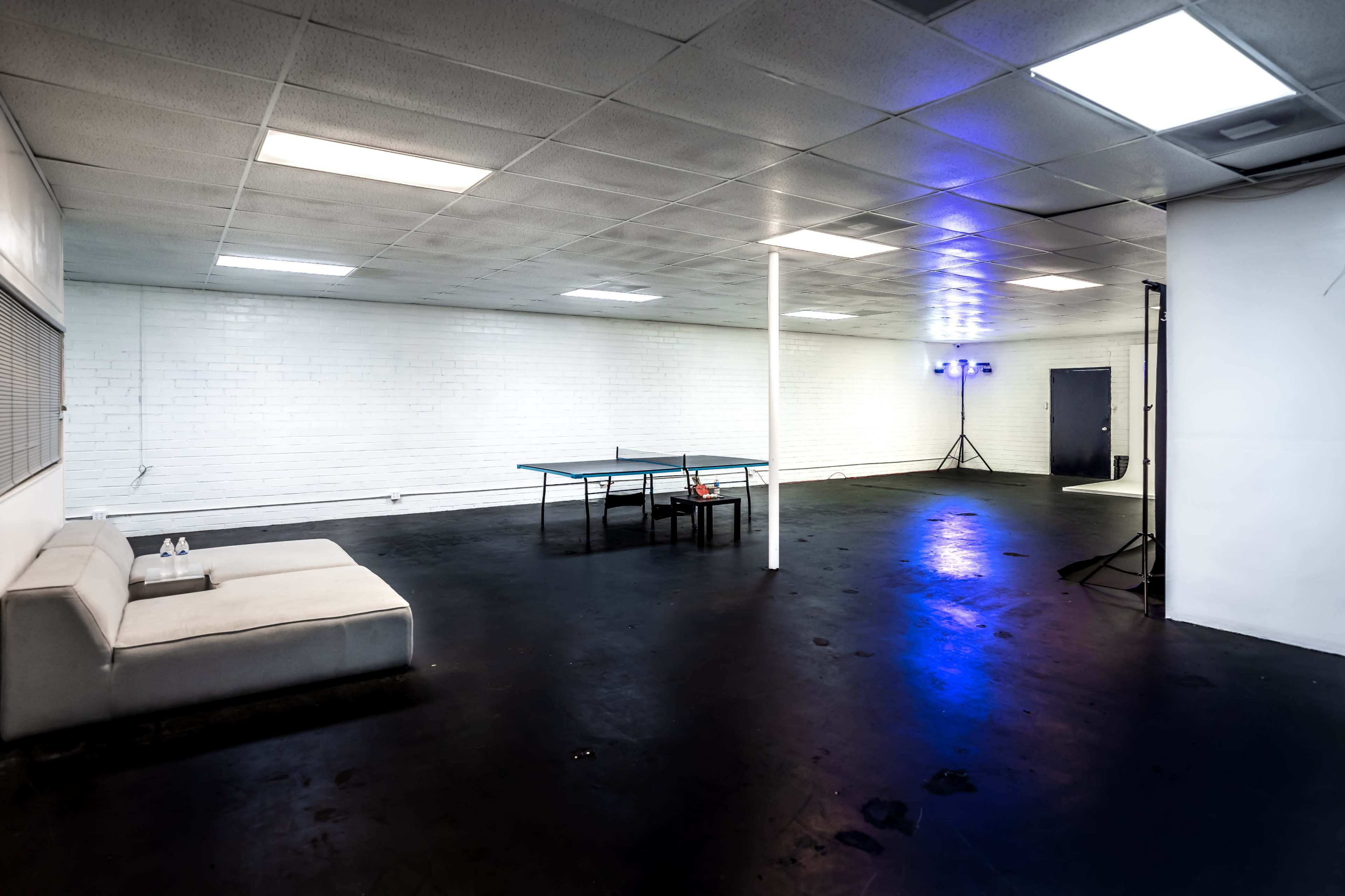 The image shows a spacious, empty room with white brick walls, a ping pong table in the center, and a sofa positioned against one wall.