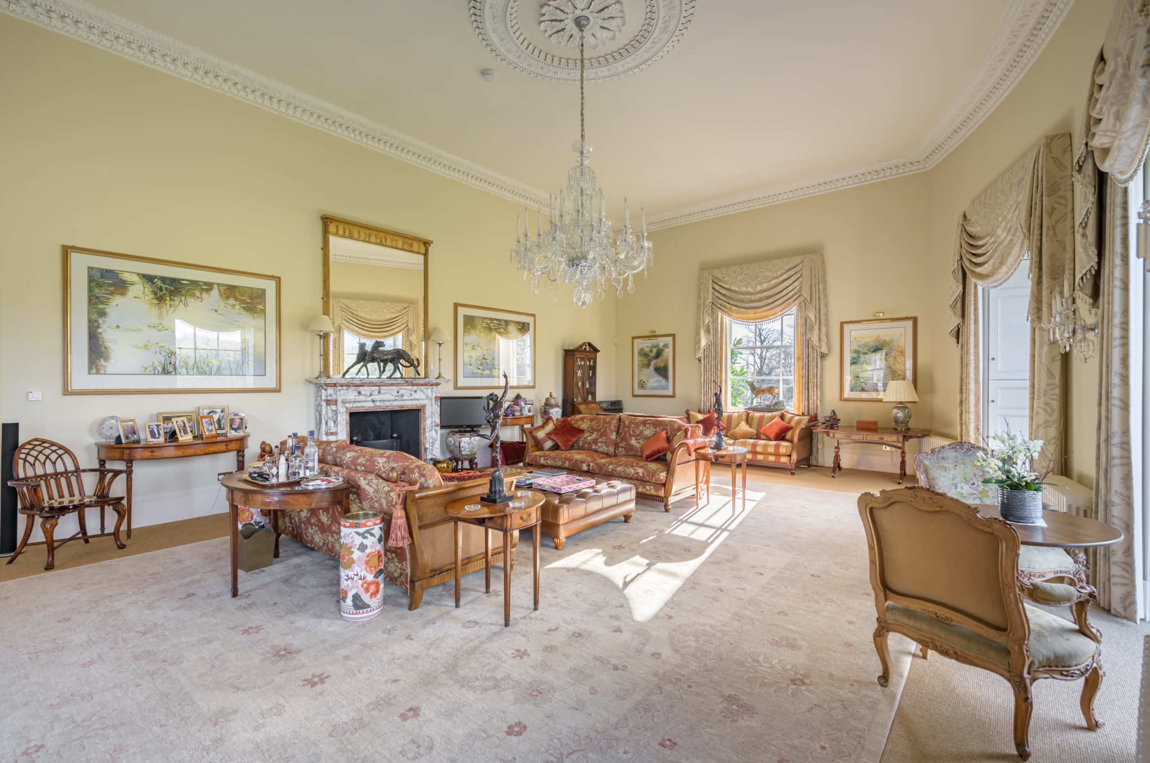 The image depicts a spacious living room furnished with ornate chairs, a chandelier, and various decorative elements, including framed art and a fireplace.