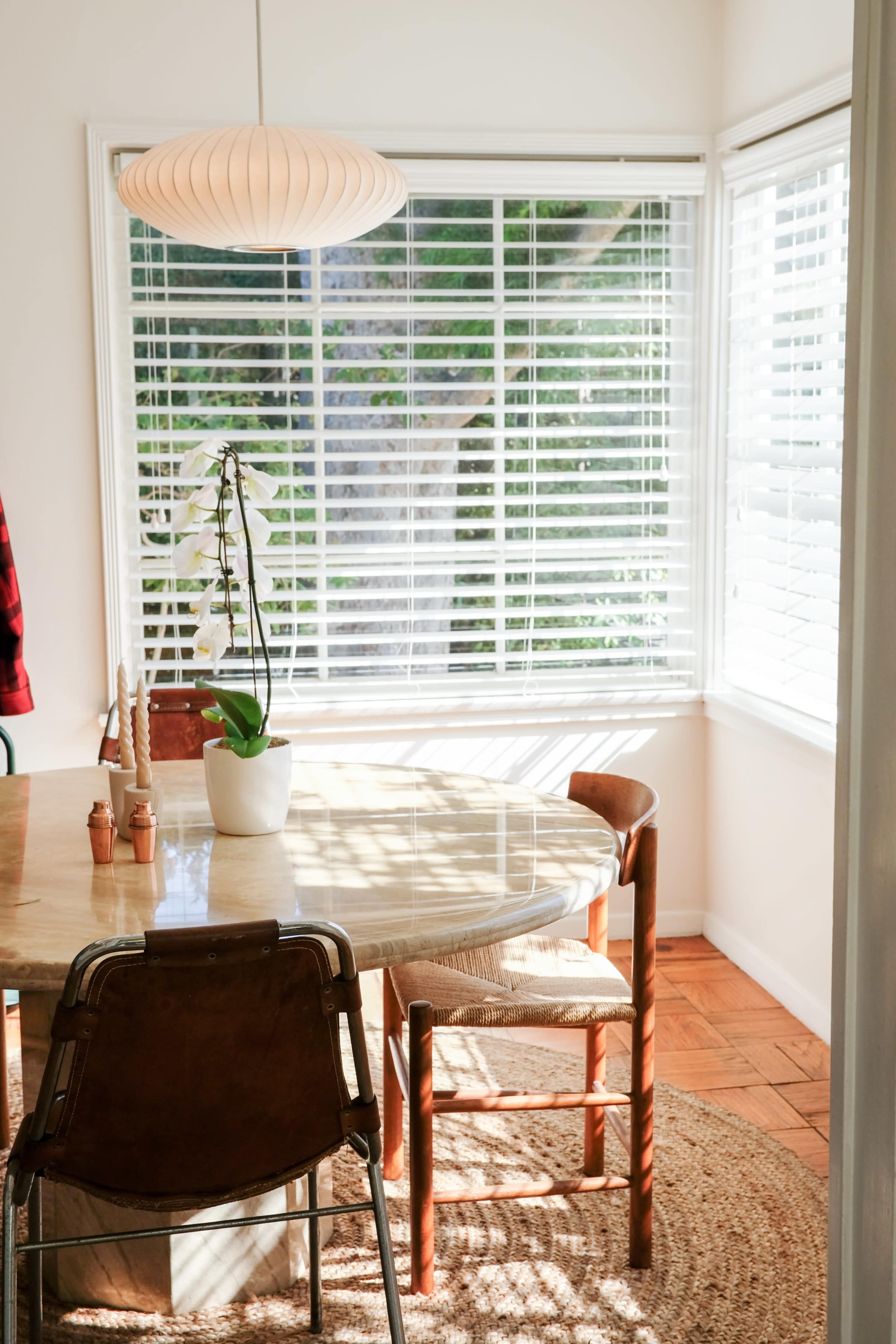 A round table with four chairs is set in a sunlit corner of a dining area, surrounded by large windows with blinds and decorated with a potted orchid.