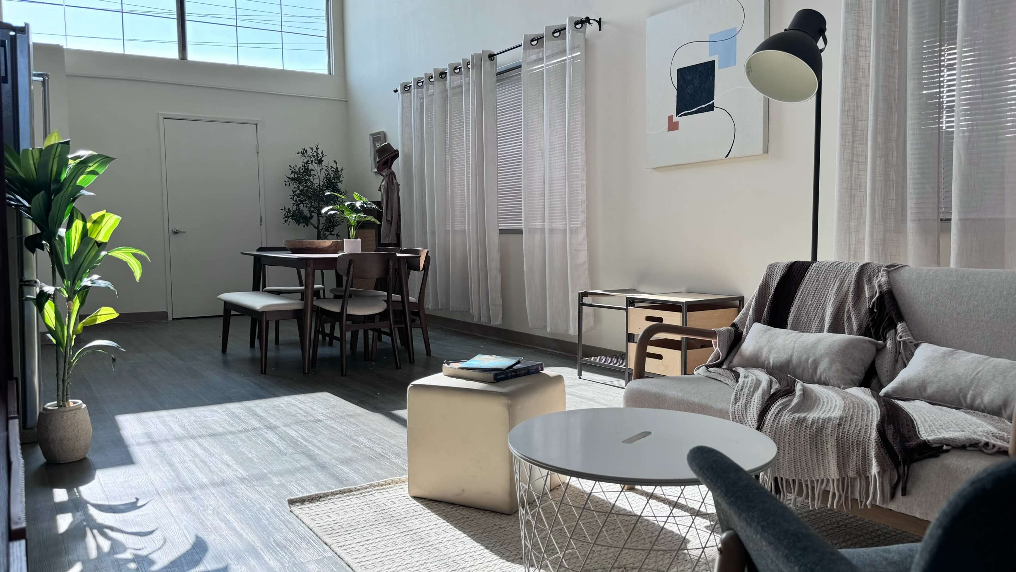A bright living space featuring a sofa, a coffee table, a dining area with chairs, and large windows letting in natural light.