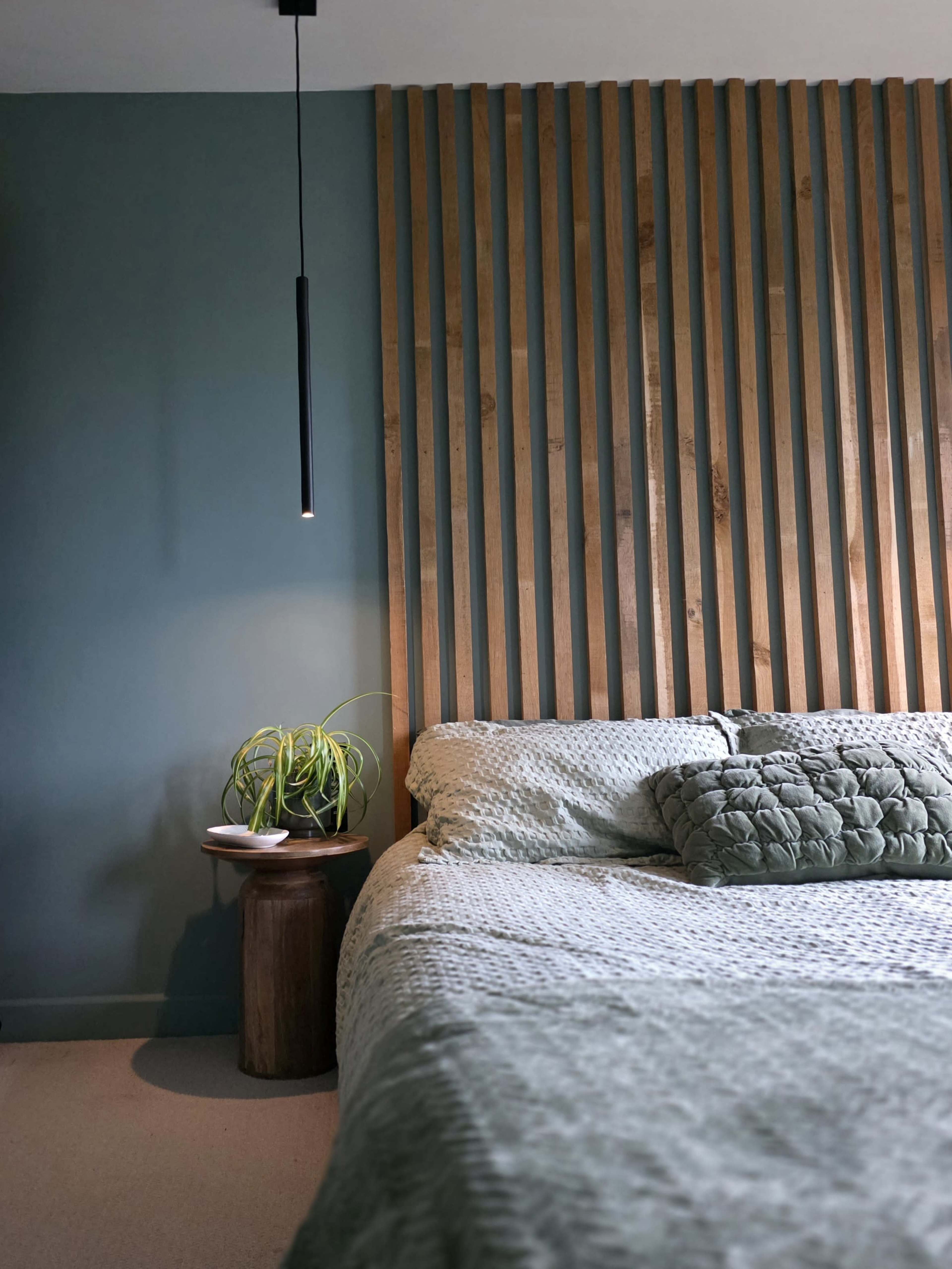 A bed with a textured blanket and decorative pillow is positioned beside a wooden bedside table with a plant, against a wall featuring vertical wooden slats.