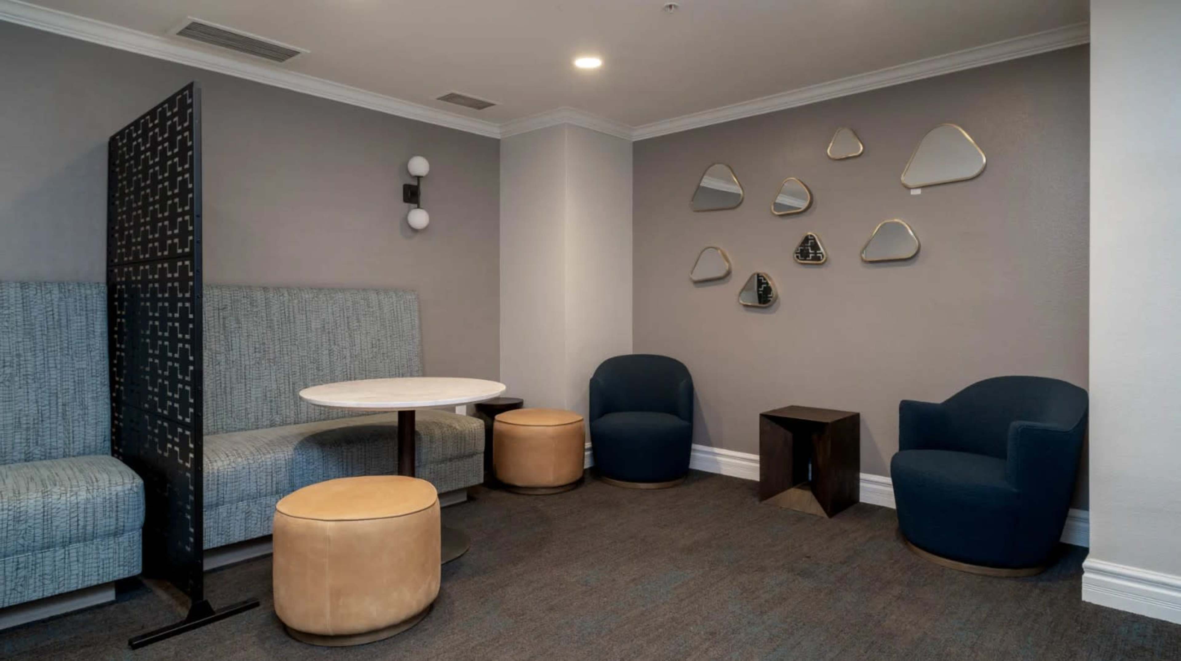 The image shows a cozy seating area featuring a round table, two blue chairs, and an upholstered bench, with abstract mirrors and a partition in the background.