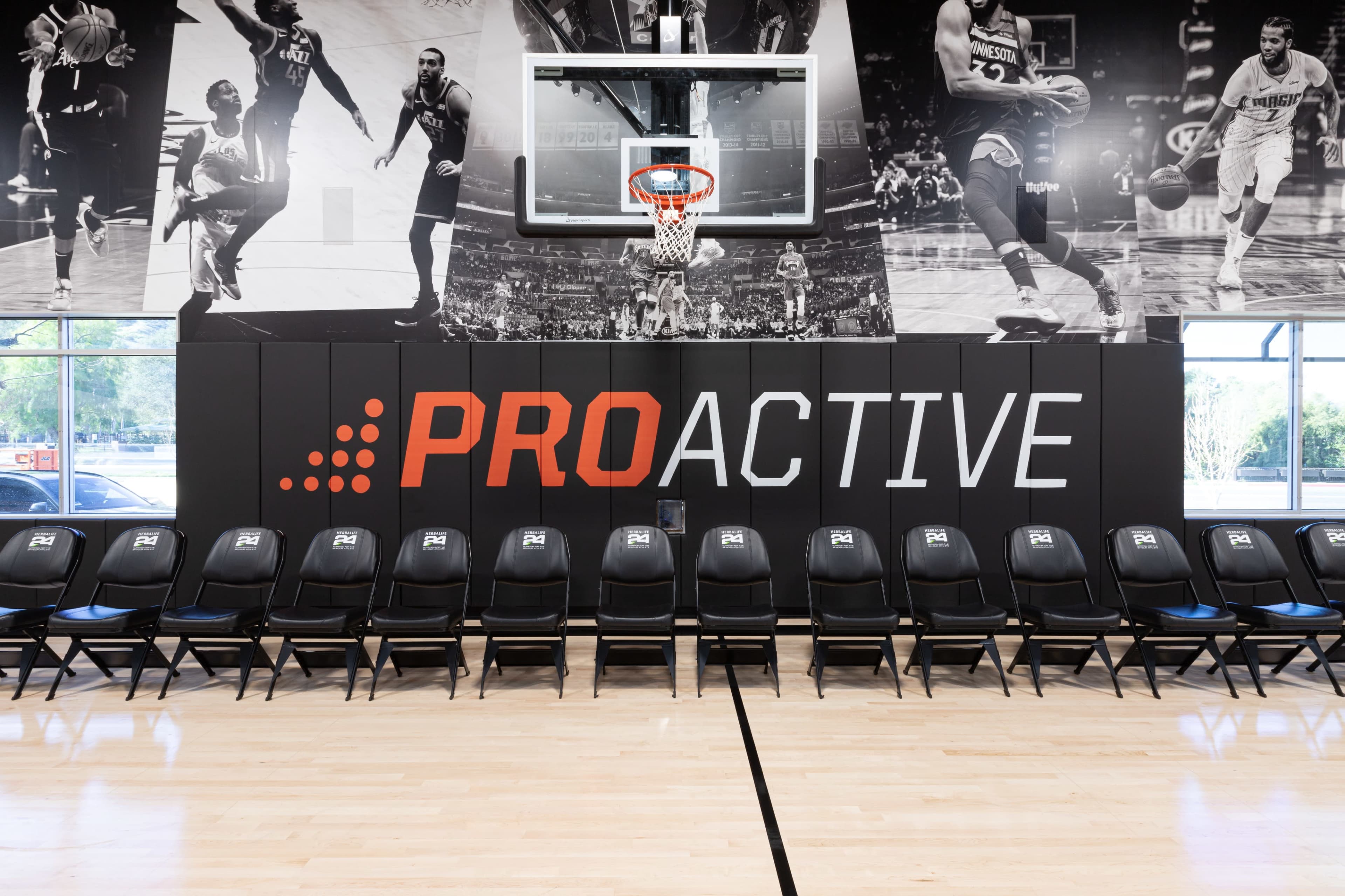 The image shows a basketball court with a large wall graphic featuring players in action behind a row of black folding chairs arranged in front of a basketball hoop, and the word "PROACTIVE" displayed prominently on the wall.