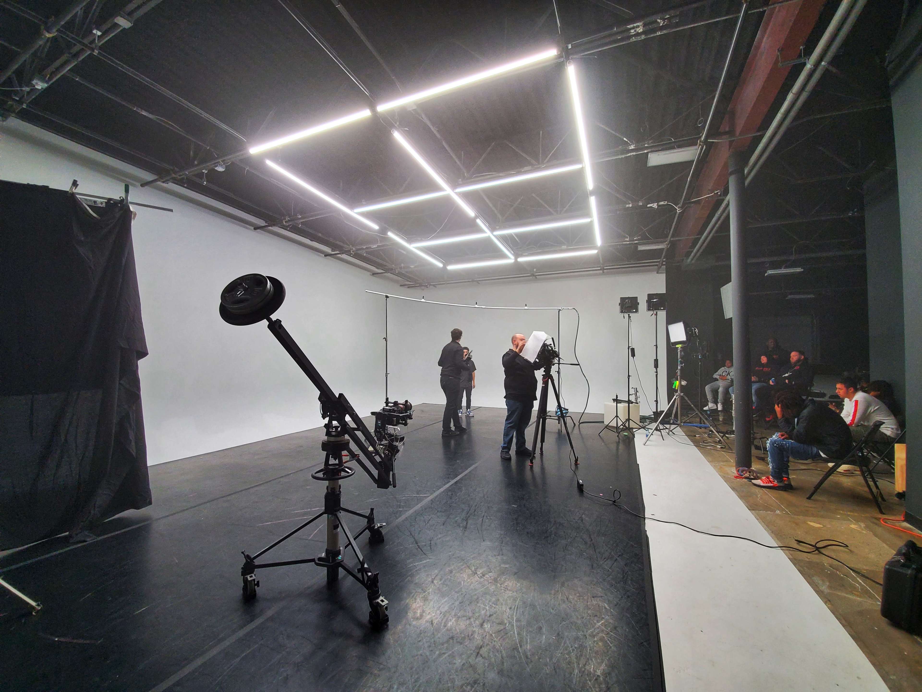 A photography studio is set up with lights, cameras, and crew members preparing for a shoot, while a group of people sits in the background.
